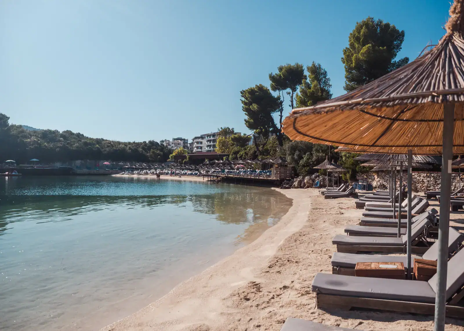 Row of sunbeds with brown mattresses and straw parasols on the right of the light colored Puerto Rico Beach, calm water with Lori Beach in the background in Ksamil on a sunny morning. 