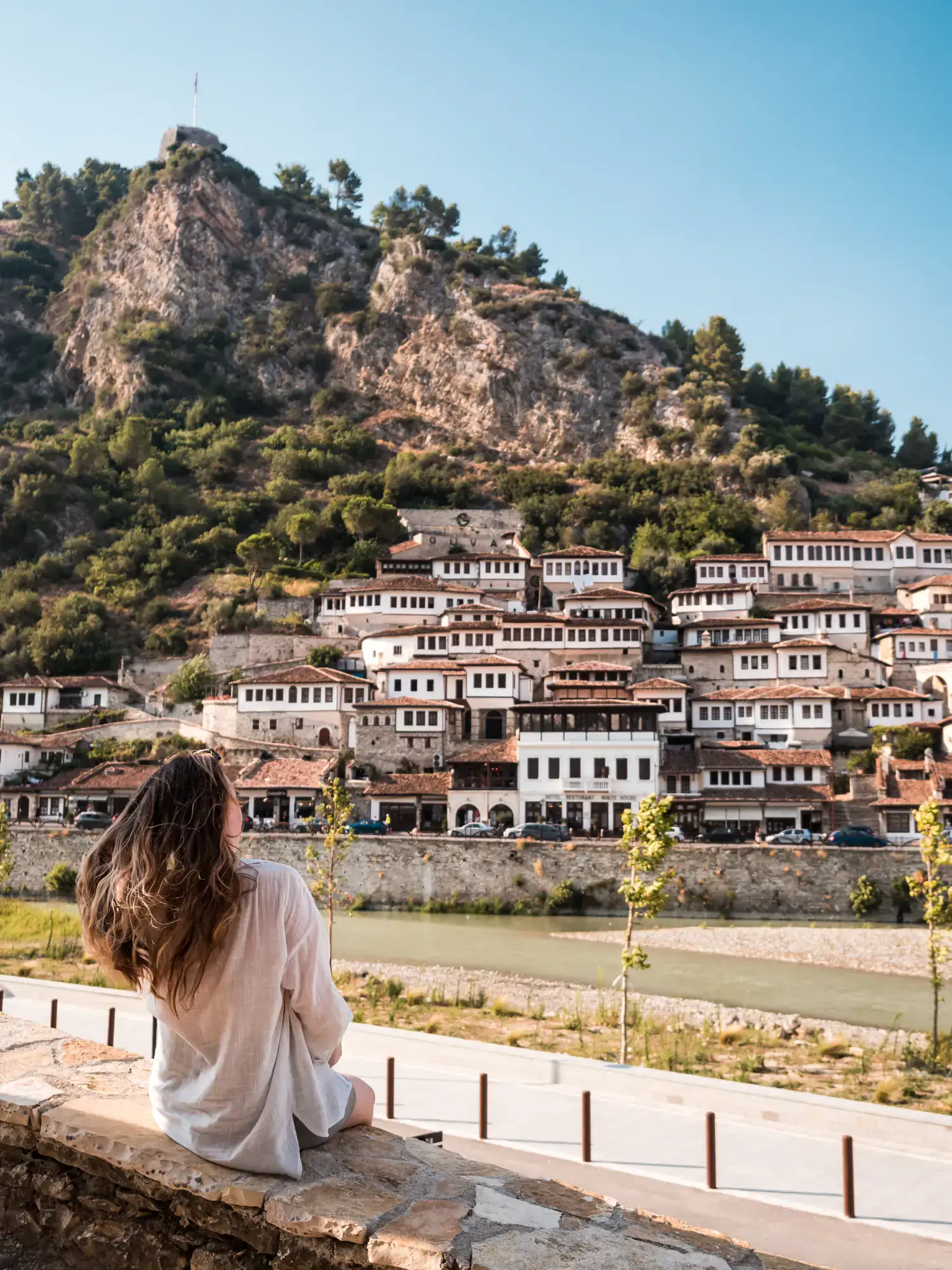 Woman wearing a white shirt sitting on a stone with her back to the camera, flicking her brown hear, looking across a shallow green river on white and brown houses with many small windows built on the hillside under a mountain in the Old Town of Berat Albania.