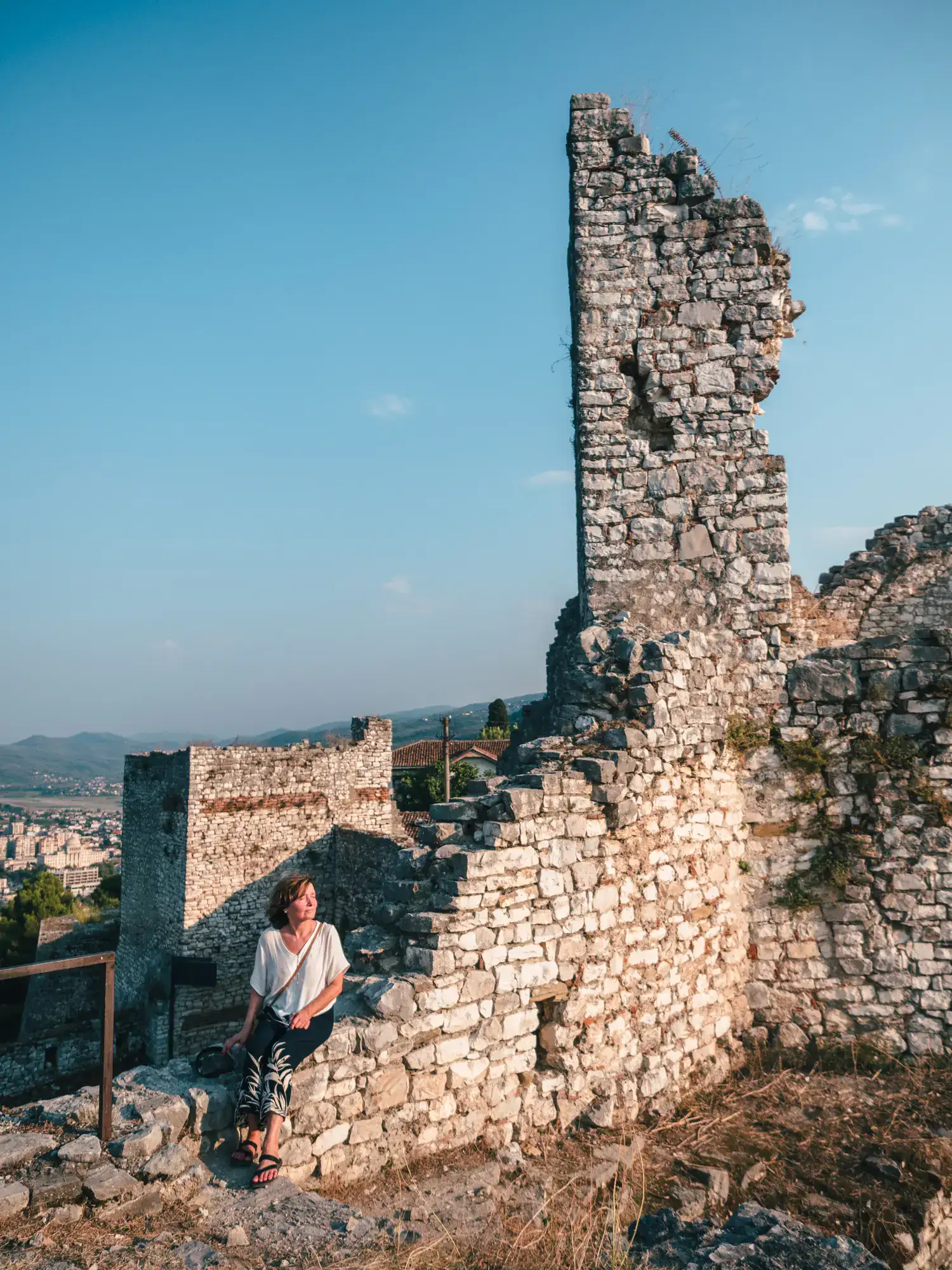 Woman wearing a white top and black pants sitting in the afternoon sun on a ruin stone wall of Berat Castle.