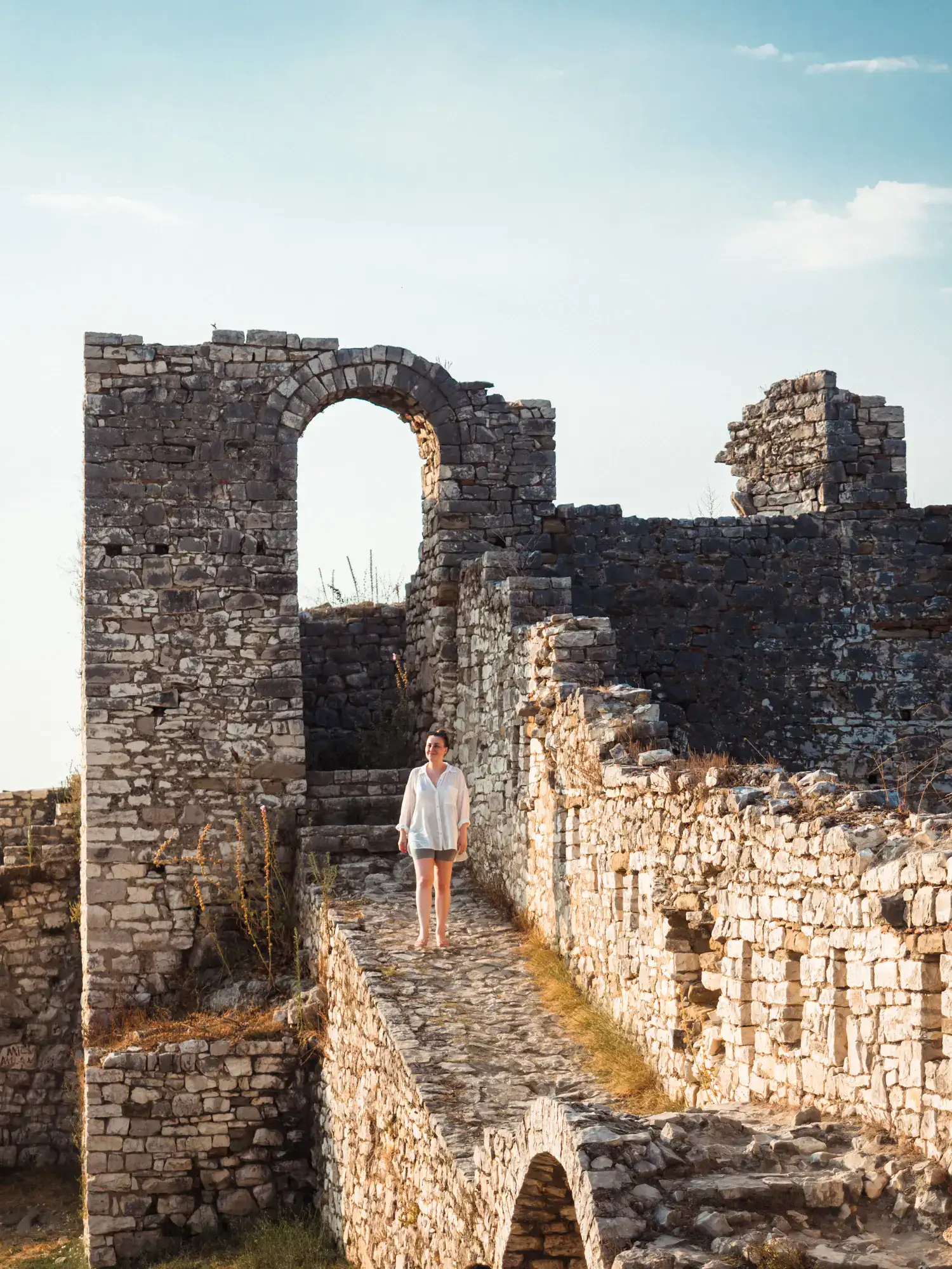 Woman wearing a white shirt and short grey shorts, walking down a cobbled walkway in the grey stone castle ruin in Berat at sunset.