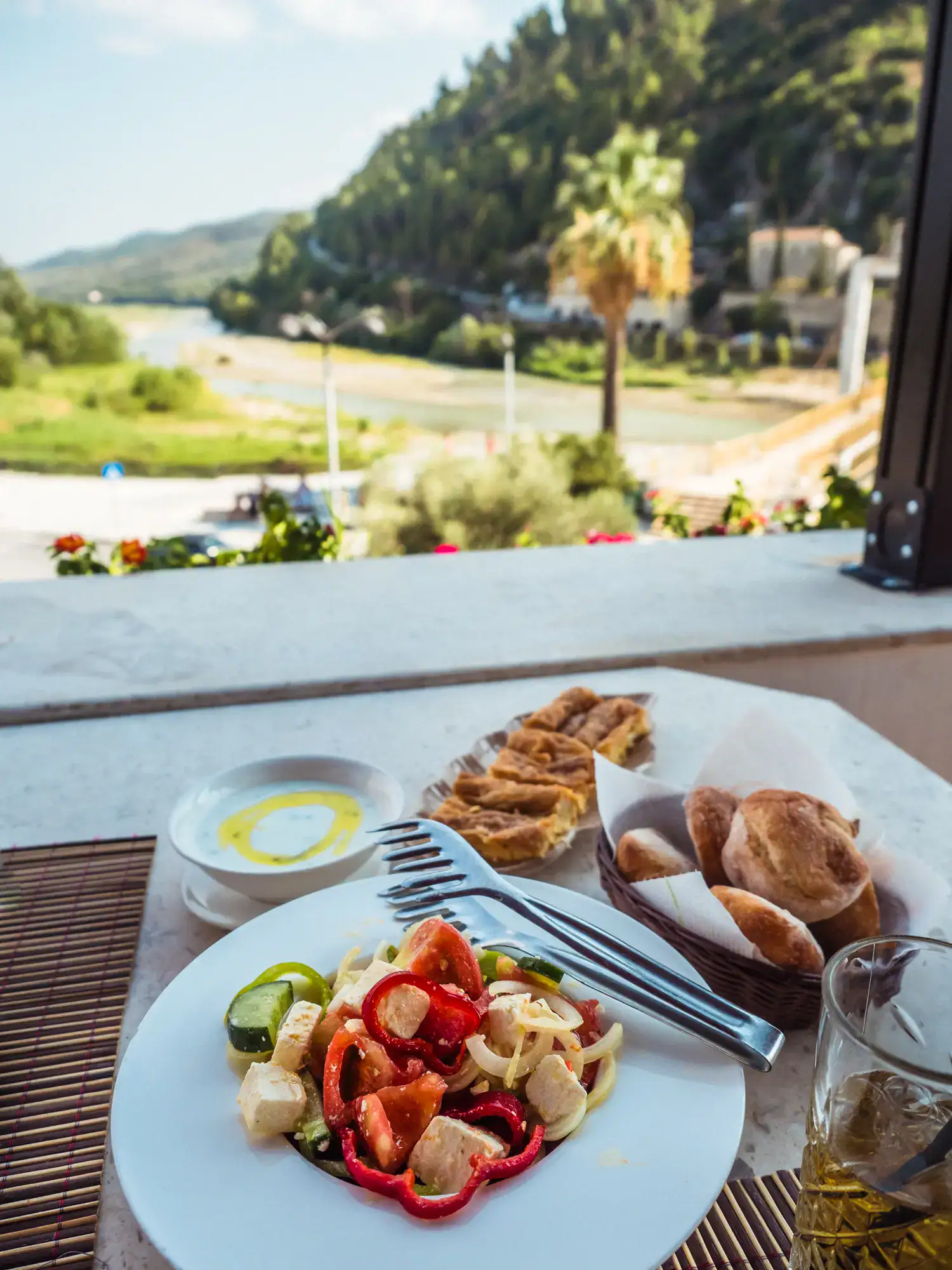 White plate with Greek Salat, white bowl with tzatziki with olive oil, bread in a basket, byrek on a white plate on a wooden table overlooking the green river and a palm tree in Berat, Albania.