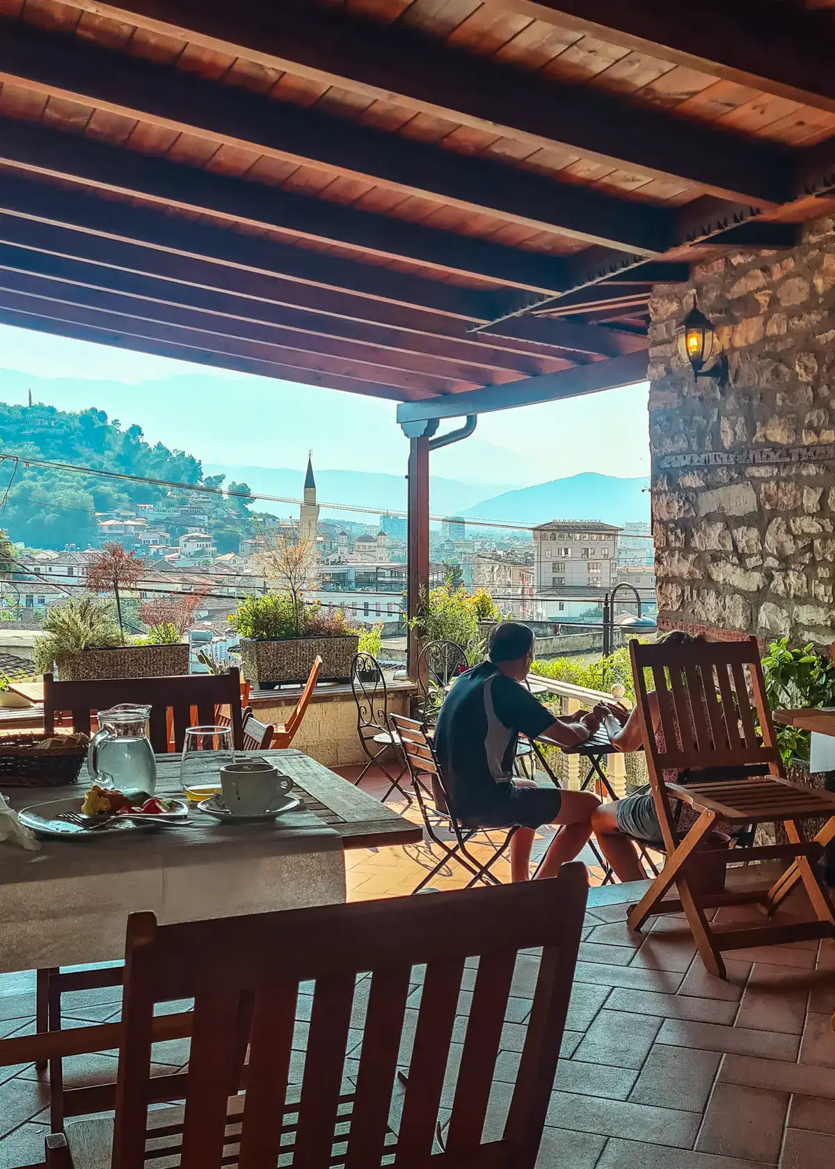 Man sitting at a wooden table in the breakfast area of Hotel Borklad under a wooden roof looking out over Berat Old Town on the tower of a mosque early in the morning.