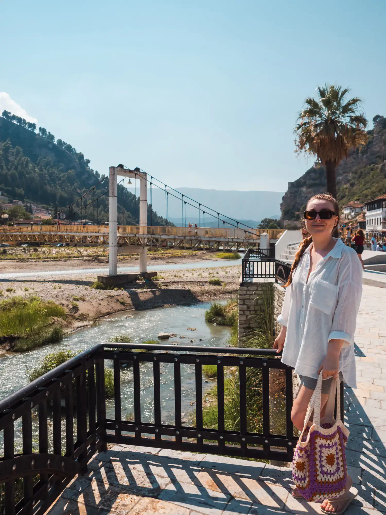 Woman wearing a white shirt, grey shorts, sunglasses, holding a crochet white and purple bag, standing by a shallow river in Berat with a white suspension bridge in the background on a sunny day. Berat Albania.