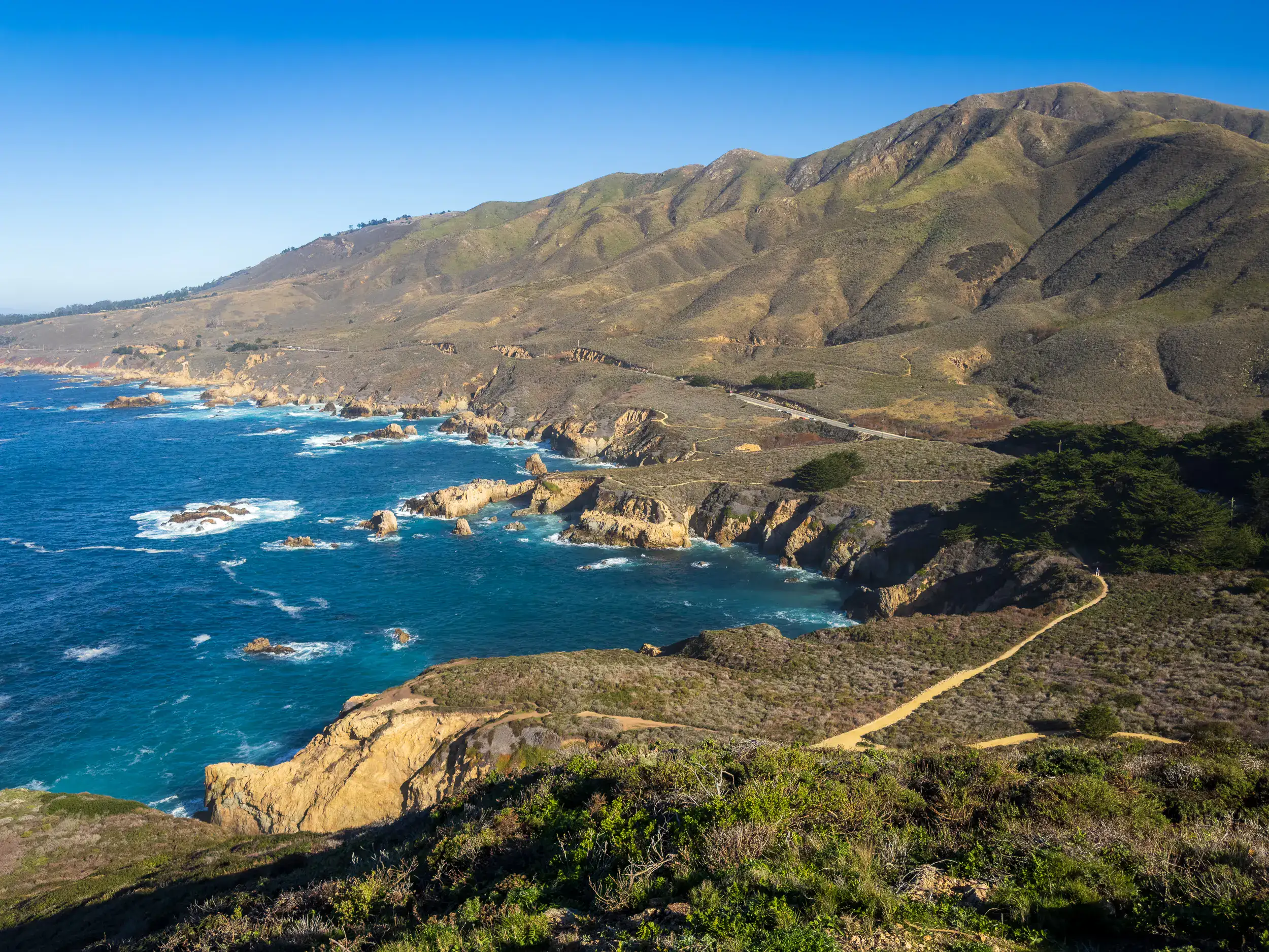 Road running along the rugged coastline of Big Sur next to deep blue ocean, on a road trip from LA.