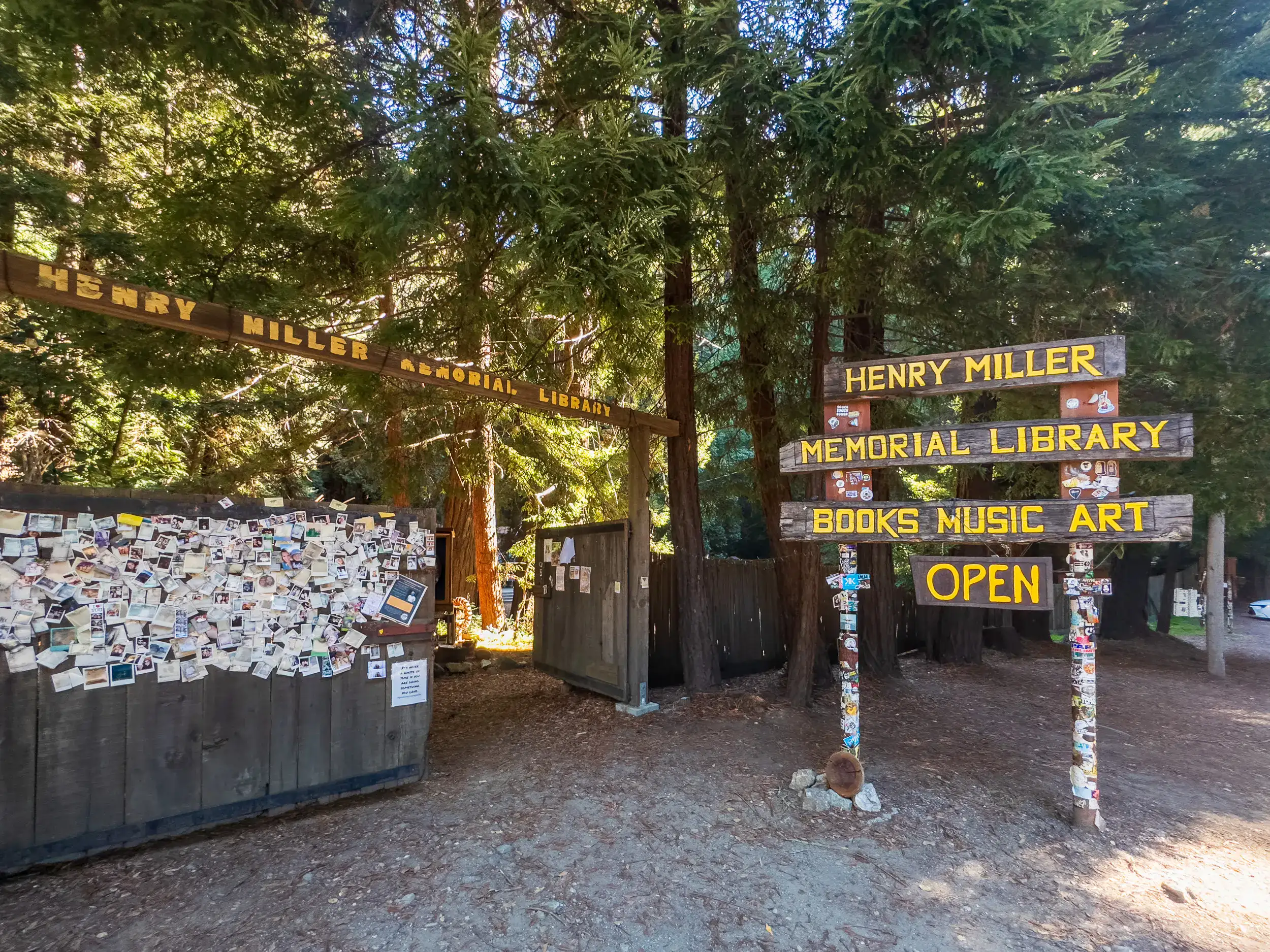 Henry Miller Memorial Library Books, Music Art, Open in yellow letters on a wooden sign outside a wooden gate and fence in the forest, on a road trip from LA to Big Sur.