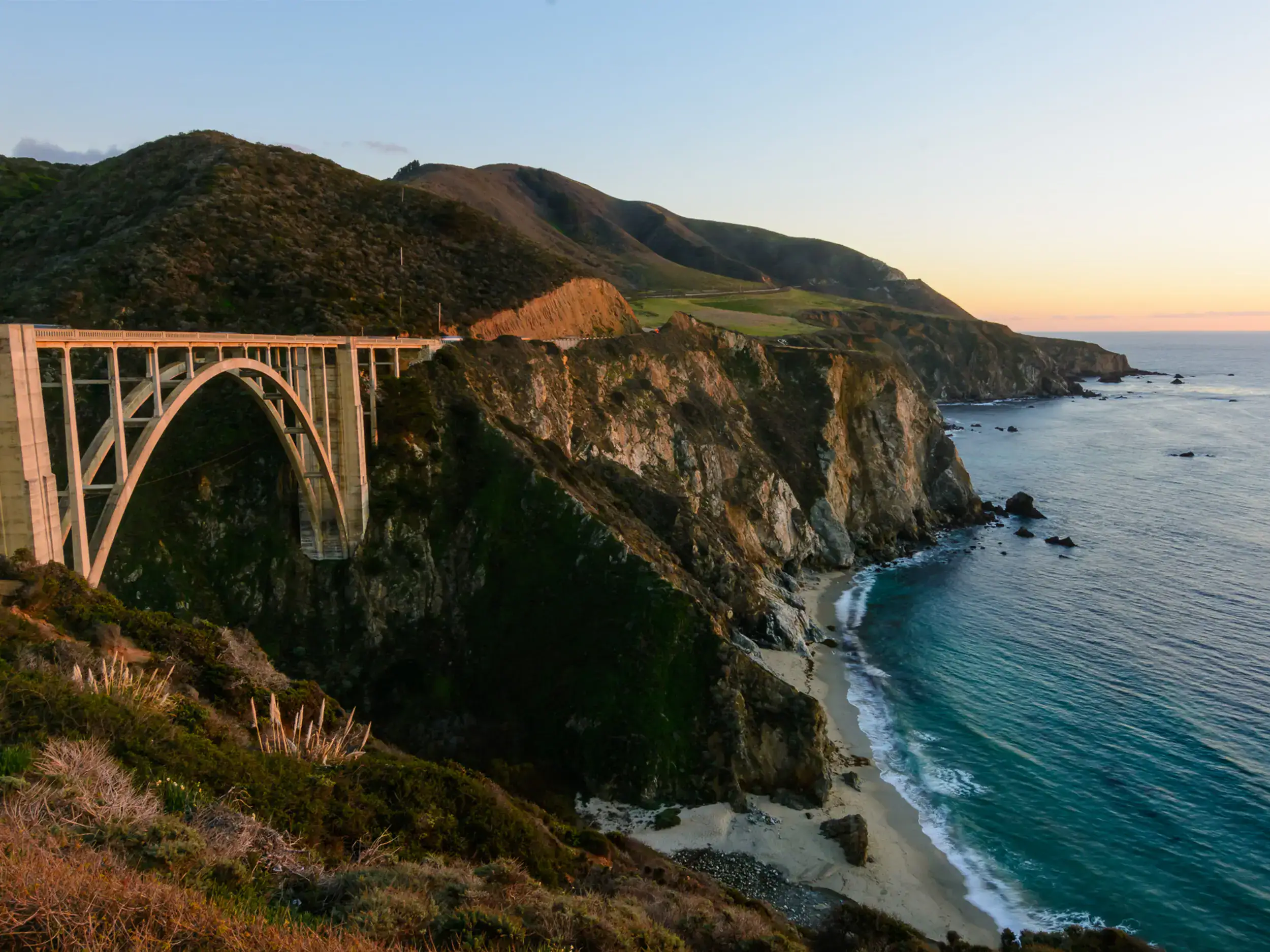 View of Bixby Bridge stretching across a mountain pass by the sea at sunset, on a road trip from Los Angeles to Big Sur.