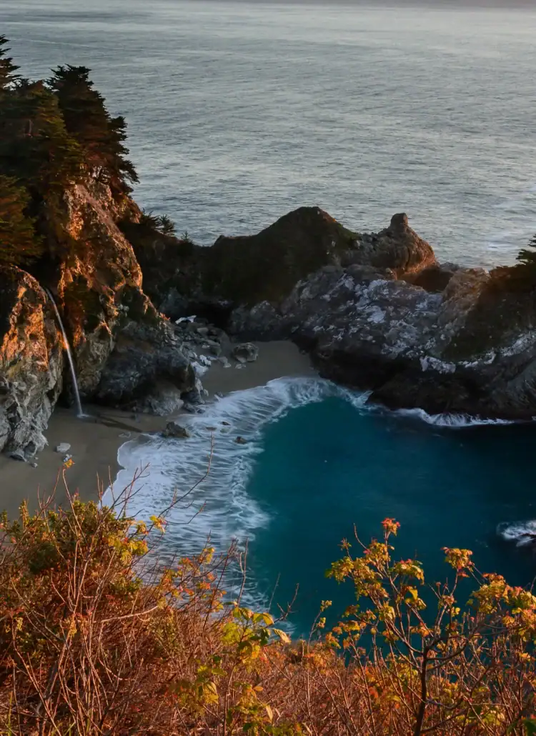Looking down, over orange brush, on a small cove with deep turquoise water surrounded by rugged stones with the small McWay Falls on a road trip from Los Angeles to Big Sur.