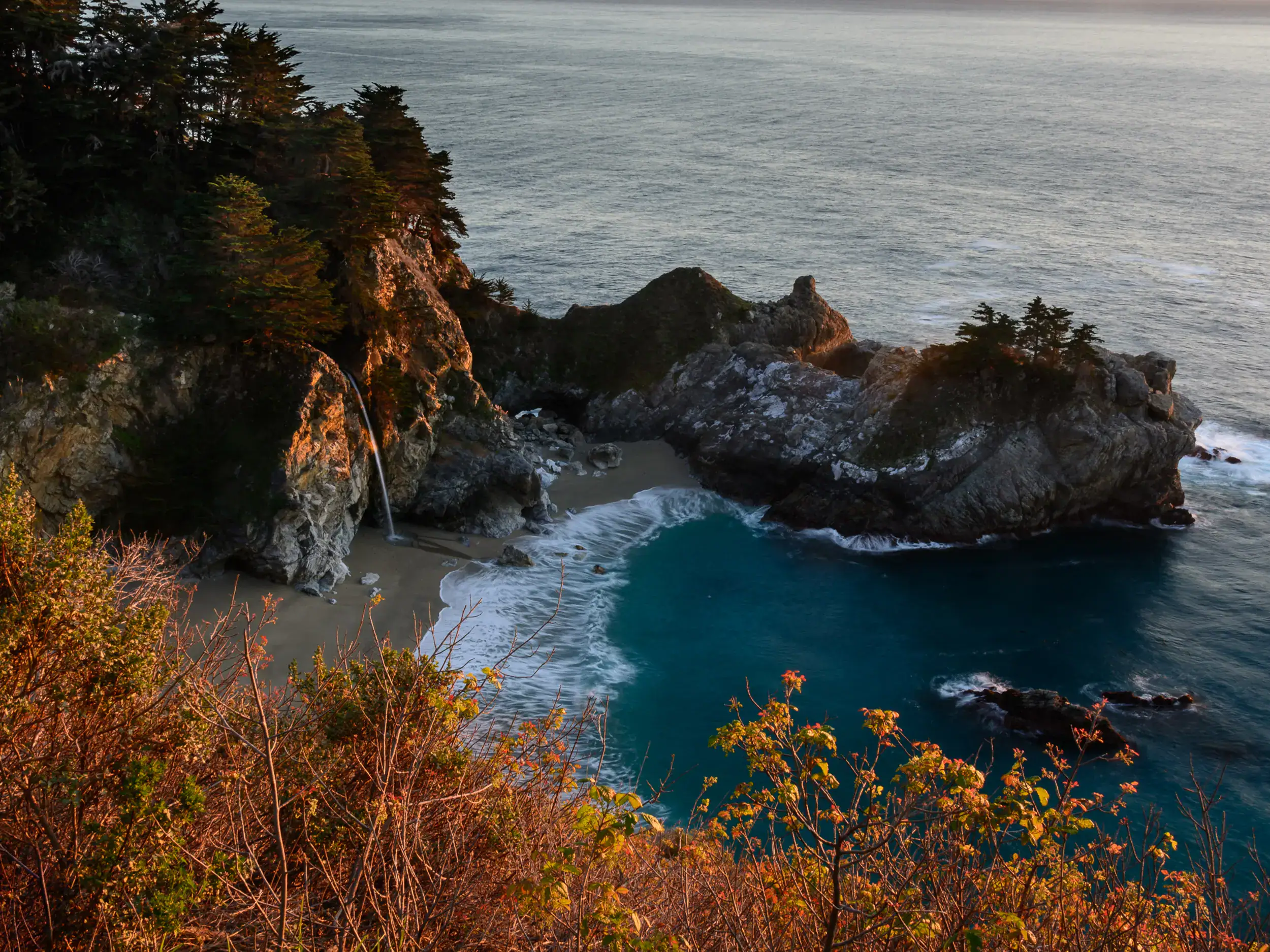 Looking down, over orange brush, on a small cove with deep turquoise water surrounded by rugged stones with the small McWay Falls on a road trip from Los Angeles to Big Sur.