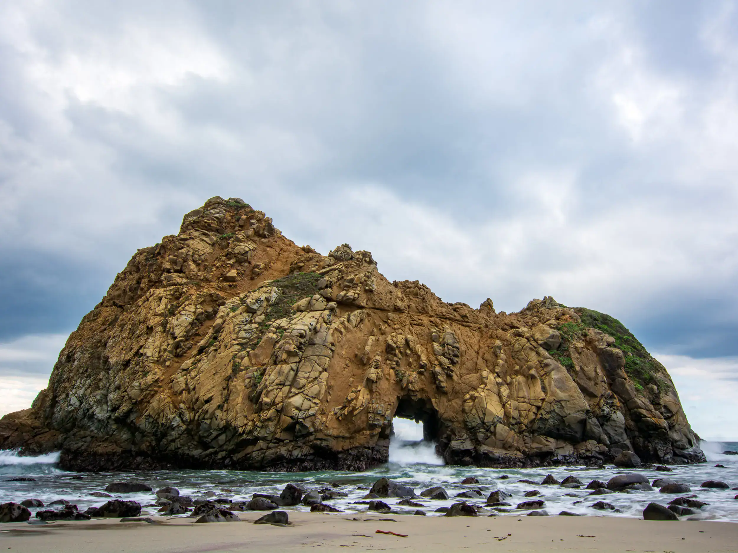Rugged stone formation with an arch in the ocean off Pfeiffer Beach with black rocks in the shallows, seen on a road trip to Big Sur from LA.