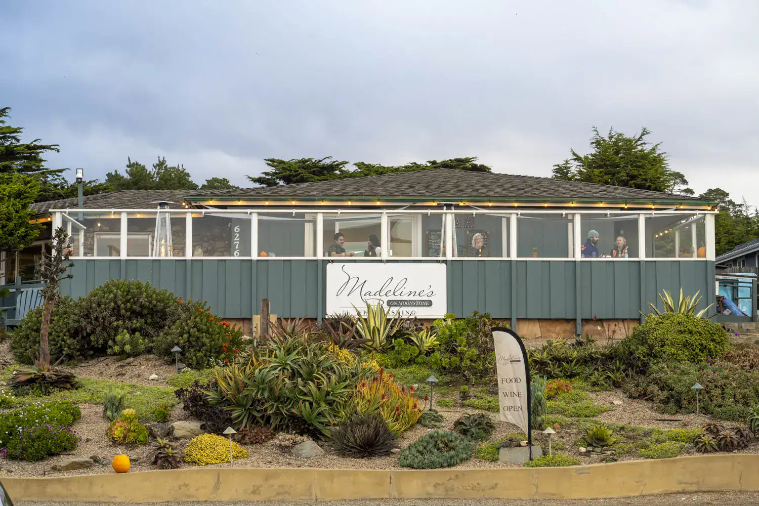 One-story wooden dark green house with a row of windows, and a white sign reading Madeleine's on Moonstone, with a flower bed in front with green brush and succulents, on a road trip from Los Angeles to Big Sur.