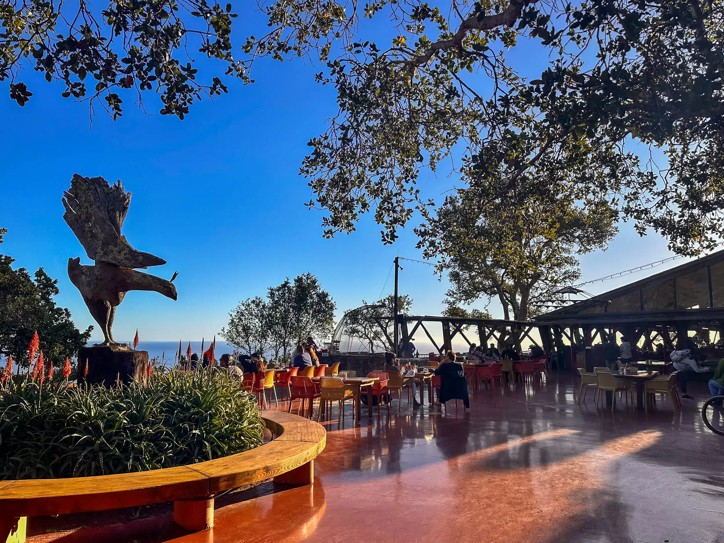 View of a brown deck, tables with yellow and red chairs, round wooden bench around greenery and a bird statue in the middle on a sunny day at Nepenthe, one of the most iconic restaurants in Big Sur.