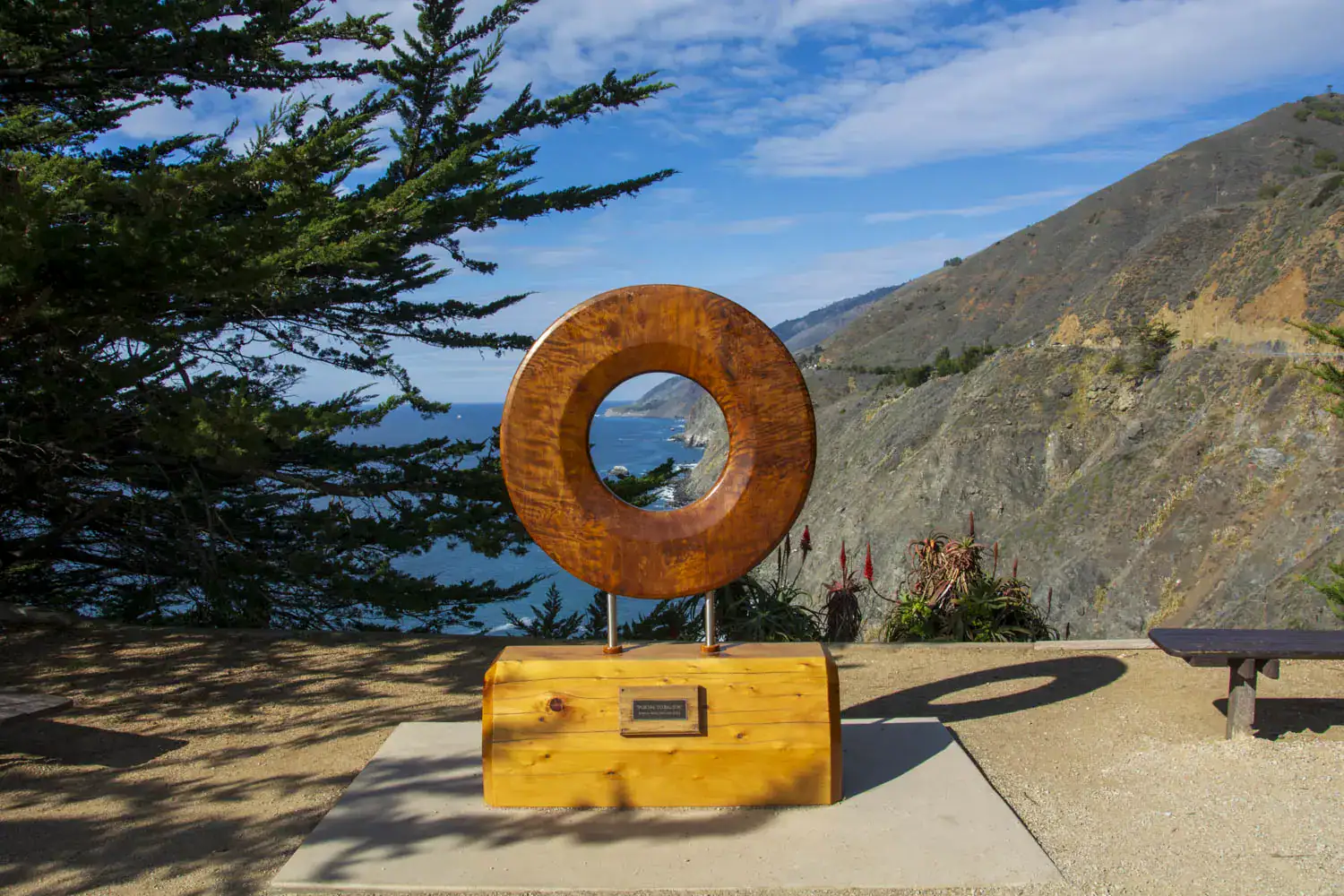 Brown wooden circle monument on a yellow wooden base on the edge of a cliff at Ragged Point Inn Lookout, seen on a road trip from Los Angeles to Big Sur.