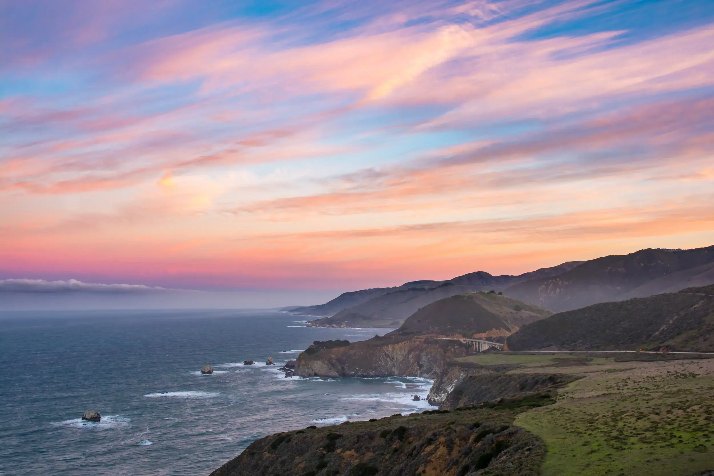 Pastel pink and orange clouds at sunrise on a blue sky, over the rugged Big Sur coast in winter on a road trip from LA.