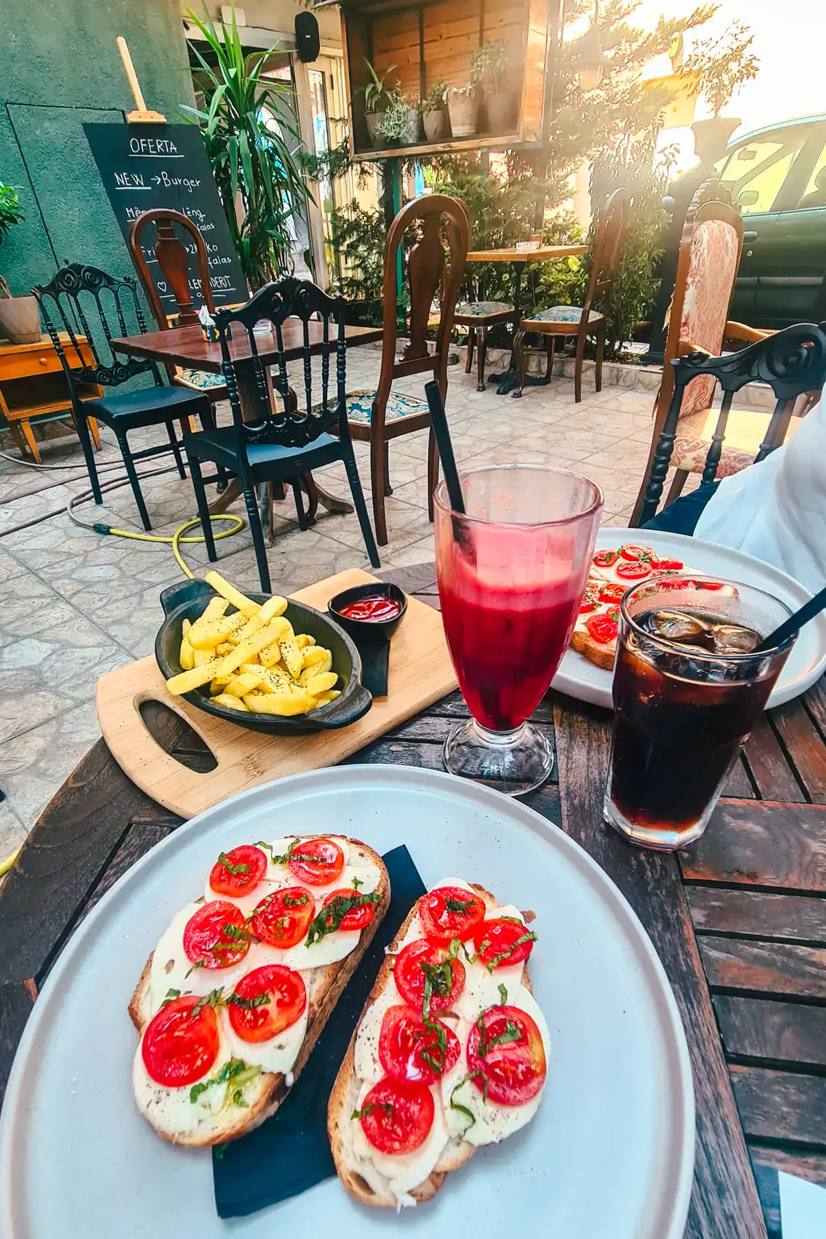 Light grey plate with two tomato and mozzarella toasts, glass of Pepsi, red juice and chips at a wooden table on the patio of Boja Bar Tattoo, one of the best cafes in Tirana, Albania.