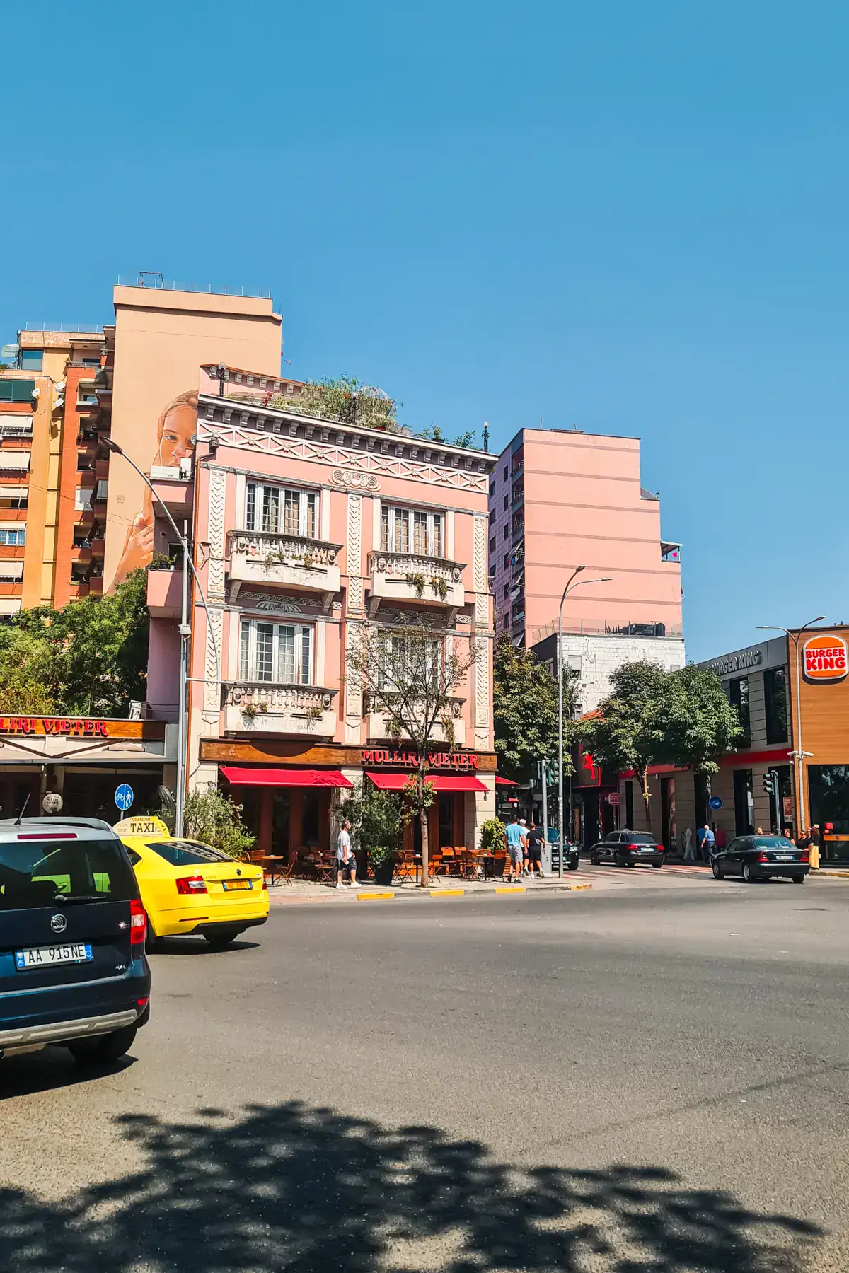 Road with a black car and yellow taxi driving on the left, light pink hotel and apartment buildings in the background on a sunny day in Tirana.