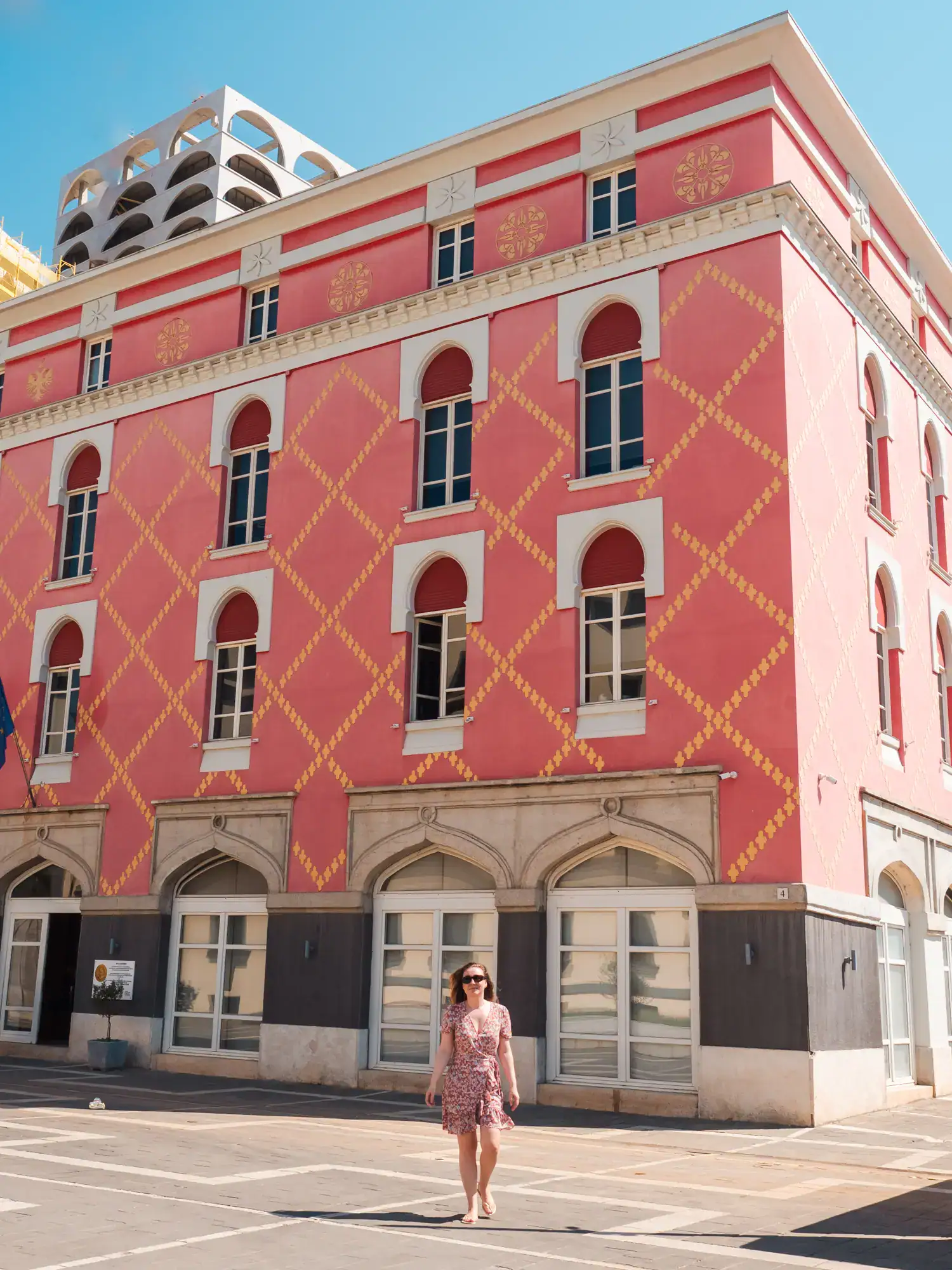 Woman in a floral dress walking in front of a vibrant pink and orange patterned building with arched windows in Tirana, Albania, on a sunny day. Is Tirana worth it?