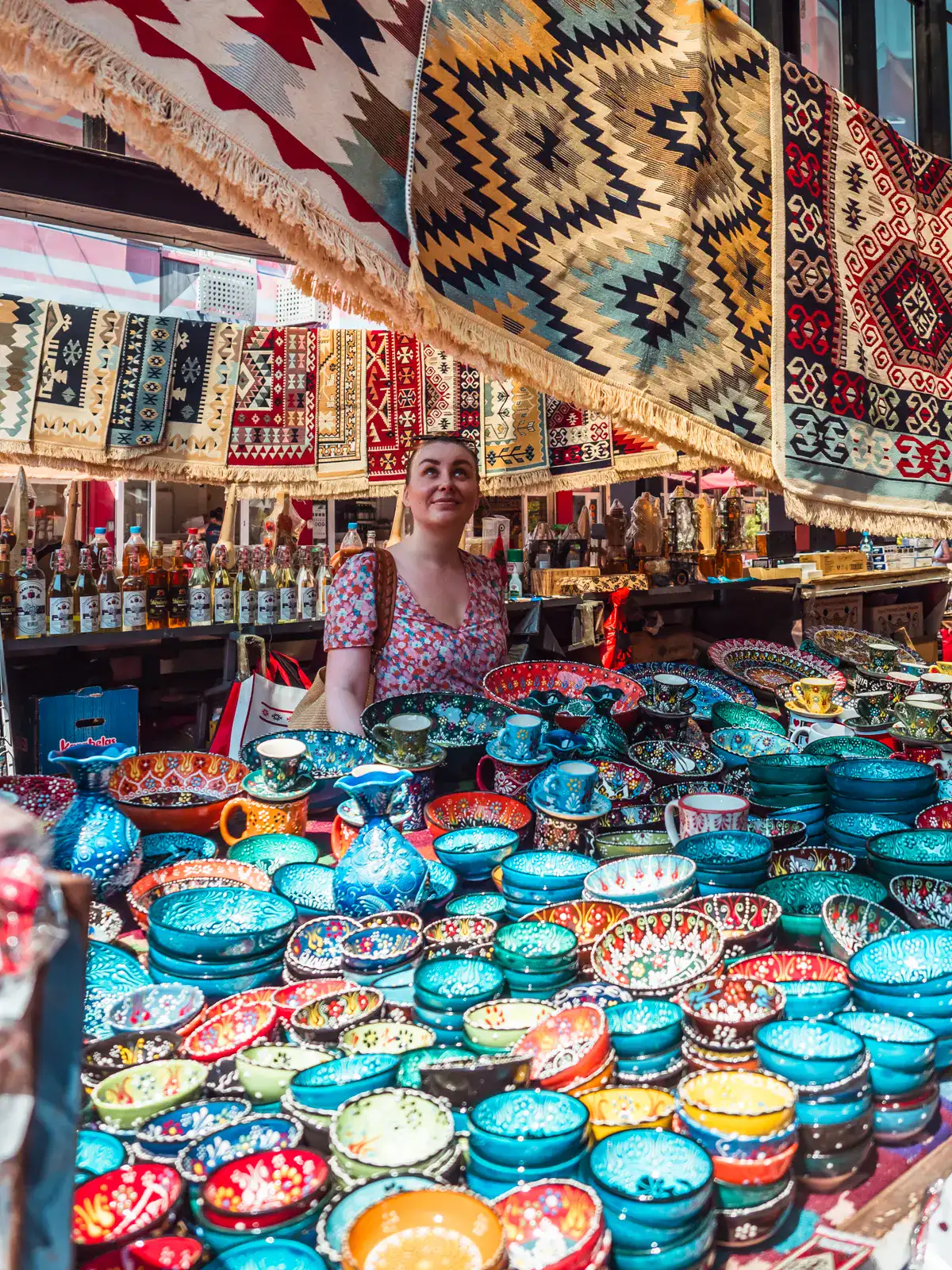 Woman wearing a pink floral dress, with her hair in a bun, looking up at patterned rugs hanging over a table with colorful pottery at The New Bazaar in Tirana, Albania.
