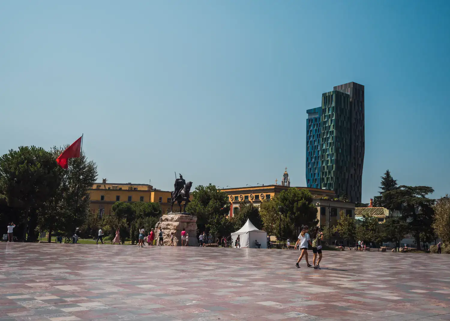 Two people walking across the vast, tiled Skanderbeg Square, with greenery and a tall geometric building in the background on the right, sunny day in Tirana.