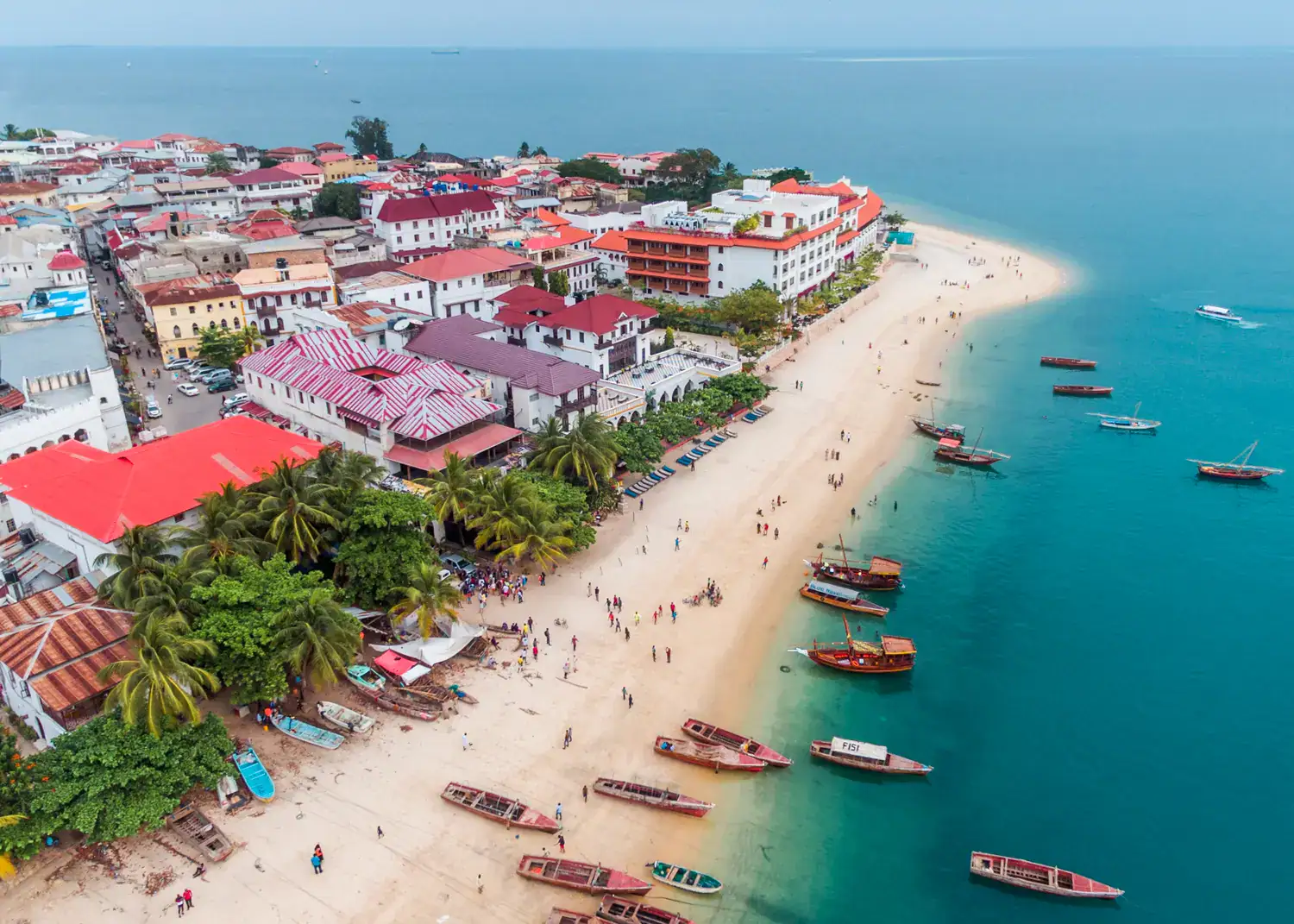 Aerial view of Stone Town Zanzibar with red, purple and orange rooftops, surrounded by greenery and a white beach with many boats, dark blue ocean.
