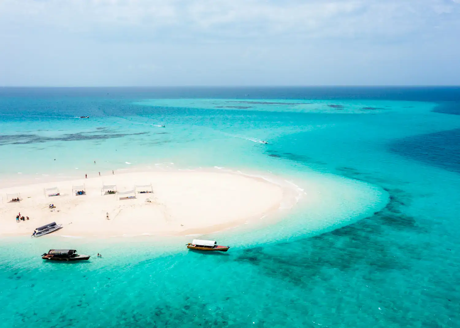 White sandbar surrounded by turquoise water with three boats, three more boats approaching, six simple tents for shade, a must on any Zanzibar itinerary.