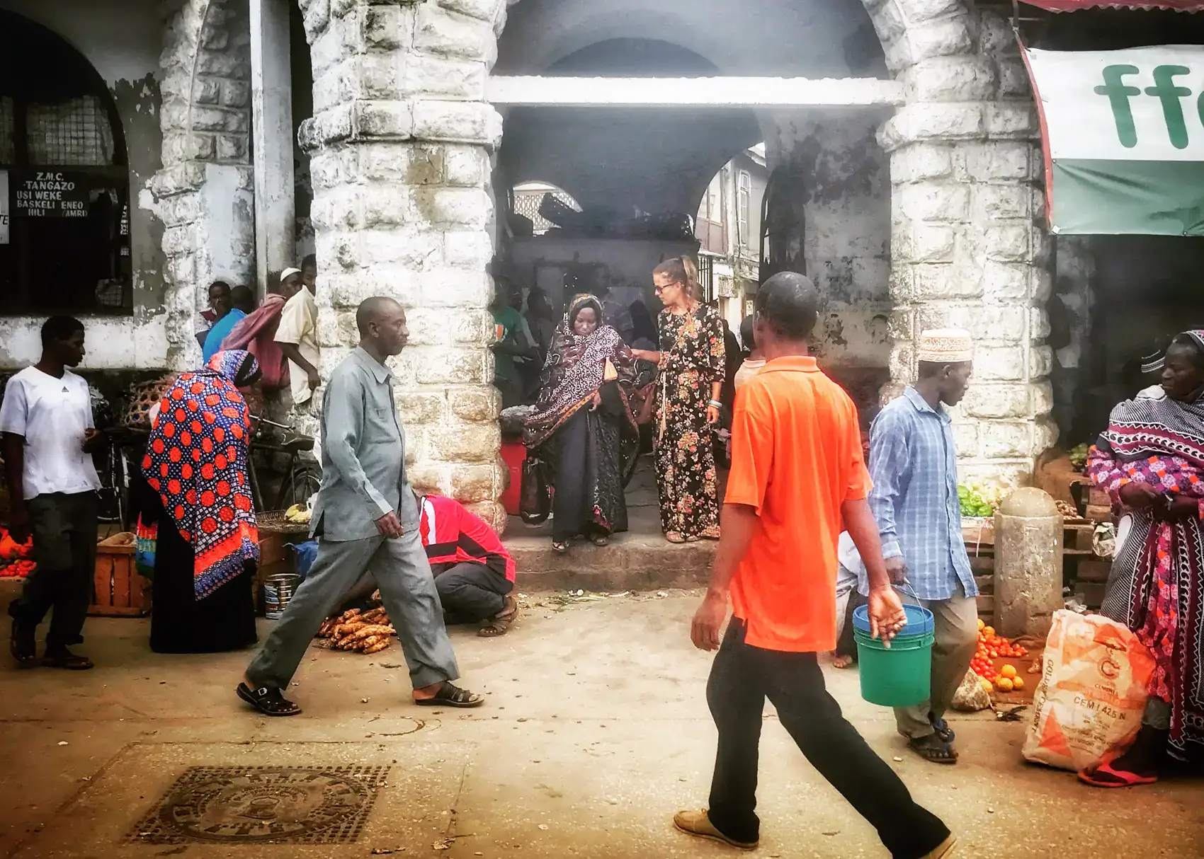 Two women wearing long patterned African style dresses walking out of a stone building in Stone Town, Zanzibar, with several men and women walking and shopping outside.