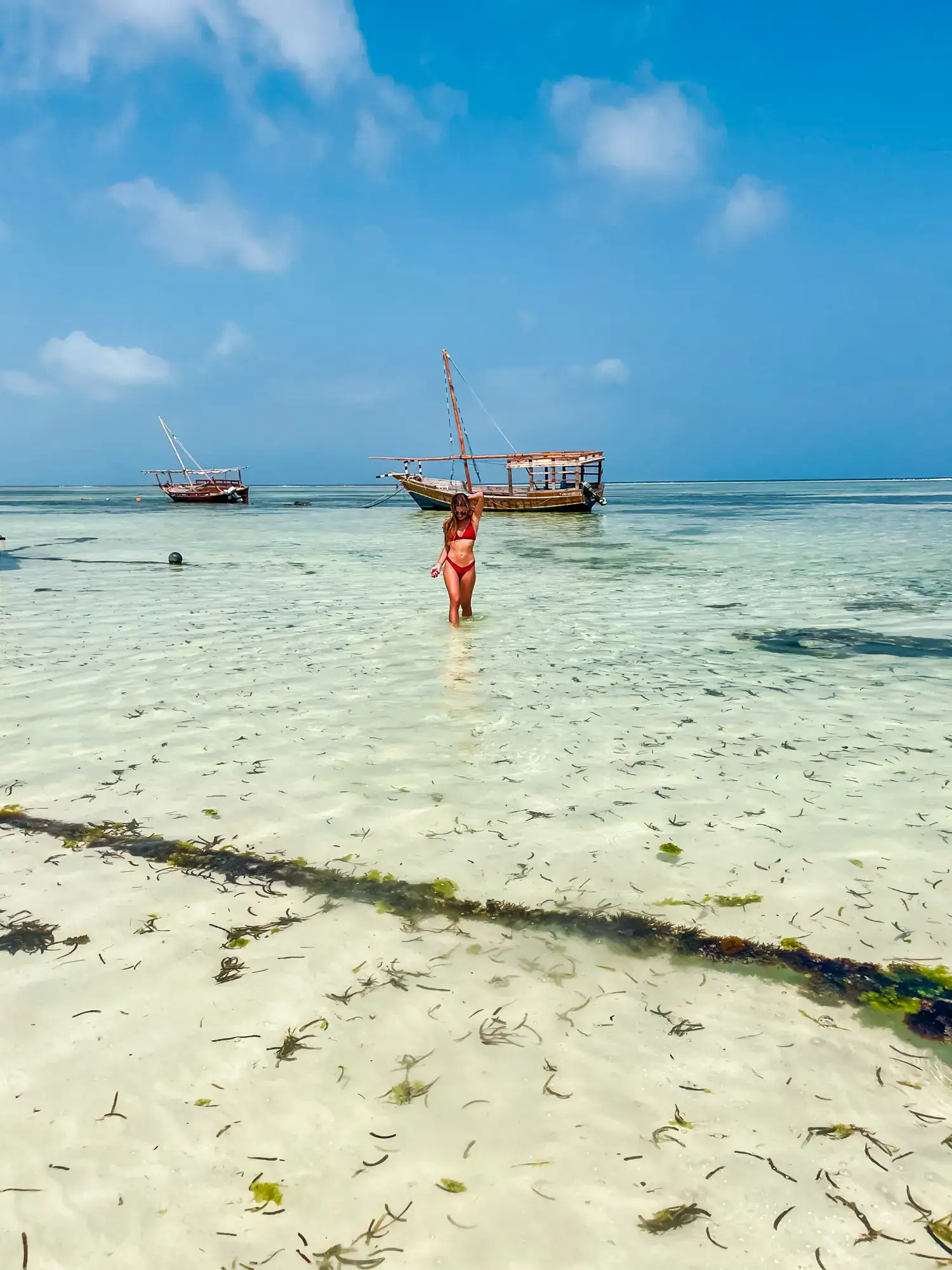 Woman with dark hear, wearing a red bikini, standing far away in shallow light green water with two boats in the background, during a sunny day in Zanzibar, Tanzania.