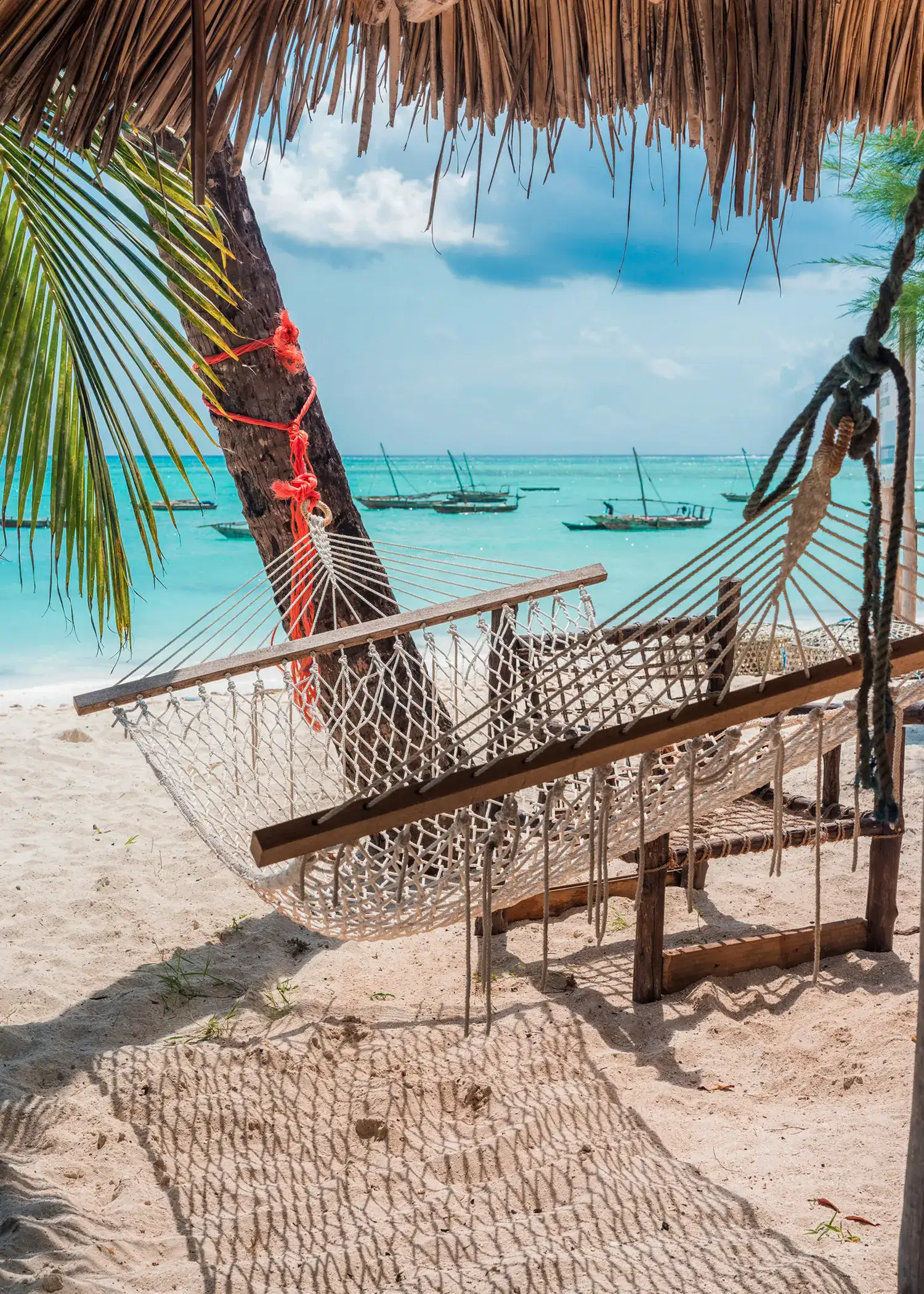 White and wooden hammock hanging from a palm tree on a white beach in Zanzibar, with turquoise ocean and boats in the background, 7-day Zanzibar itinerary.