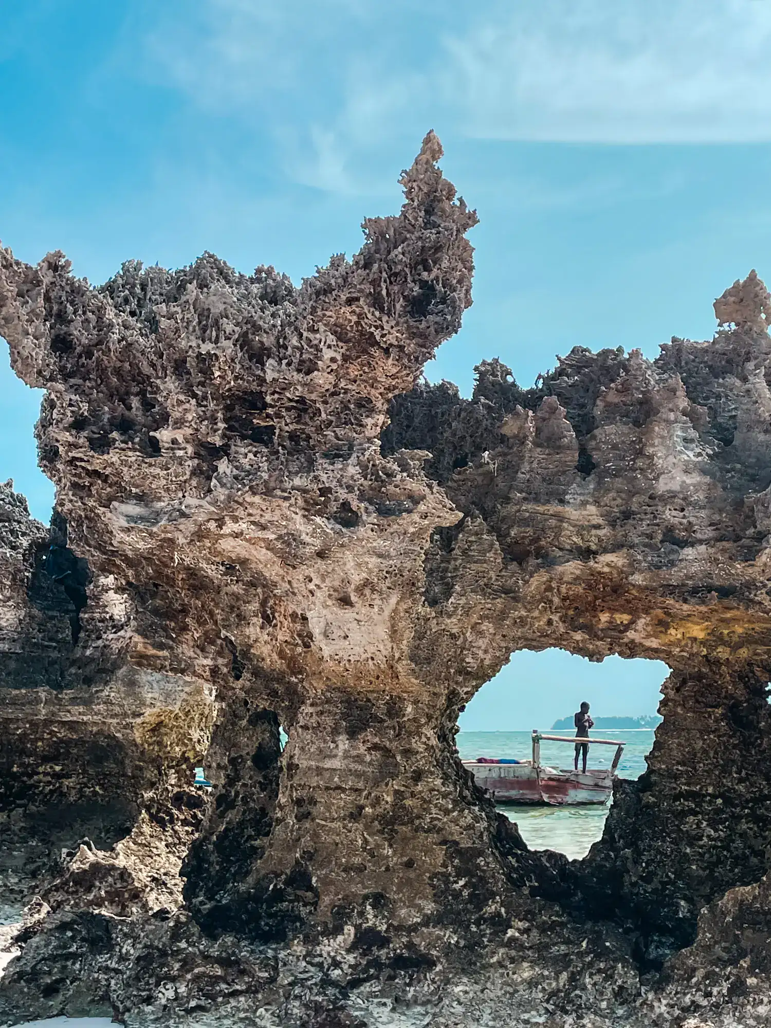 Sun is shining, boy standing in a small wooden boat seen through a round whole in a tall, coral like stone structure on one of the best beaches in Zanzibar.