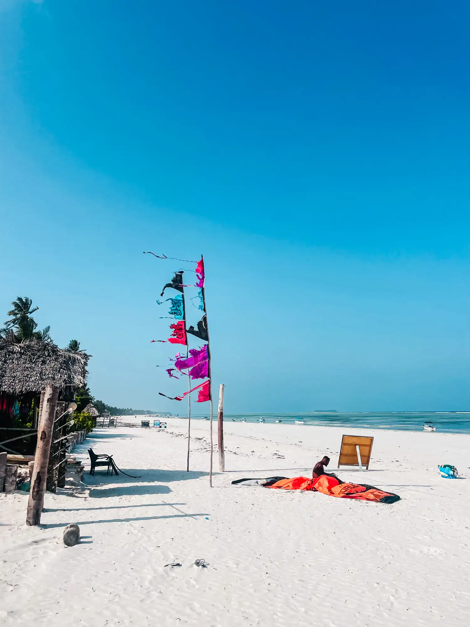 Man sitting with a orange parafoil on a white beach, two sticks with purple, blue and pink flags and a beach shack in the background, the perfect Zanzibar itinerary.