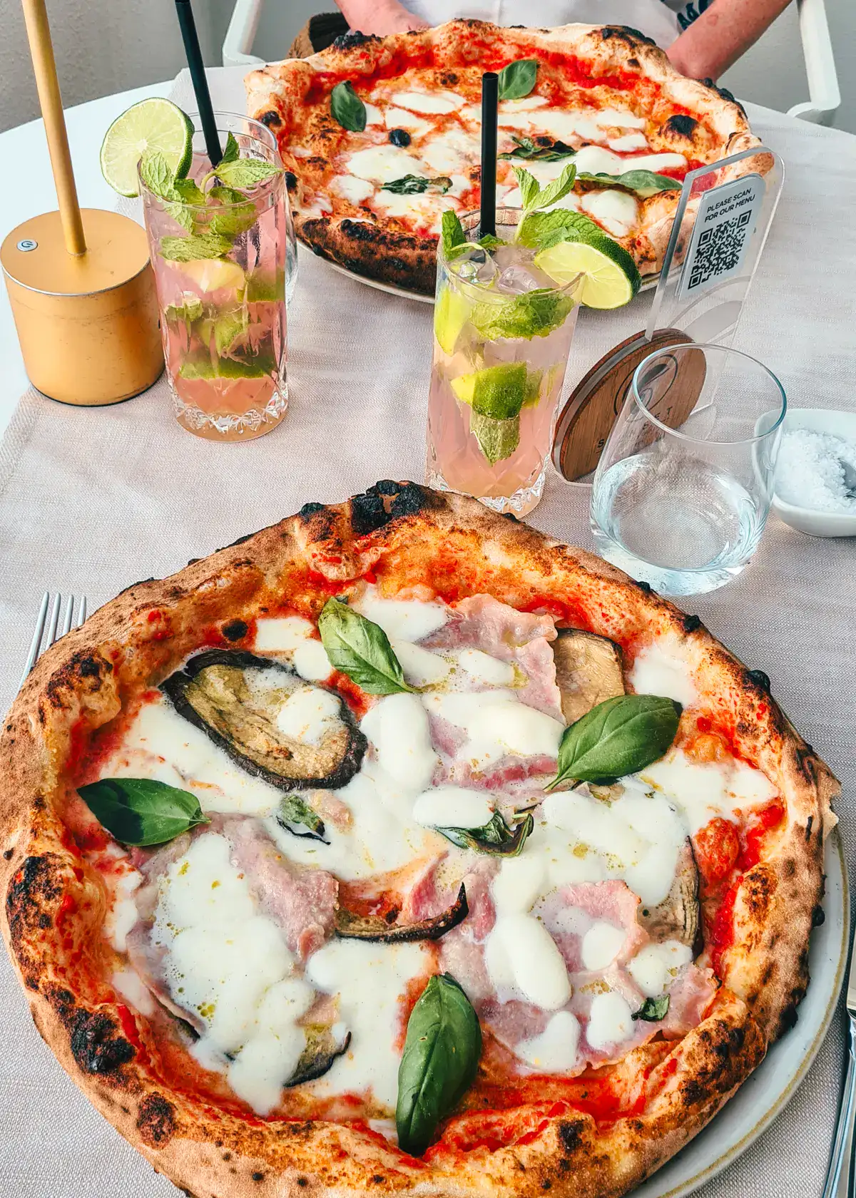 Pizza Capricciosa with fresh basil on a white table with a light pink cocktail with limes in the background at Sunset Beach Club Tropea.