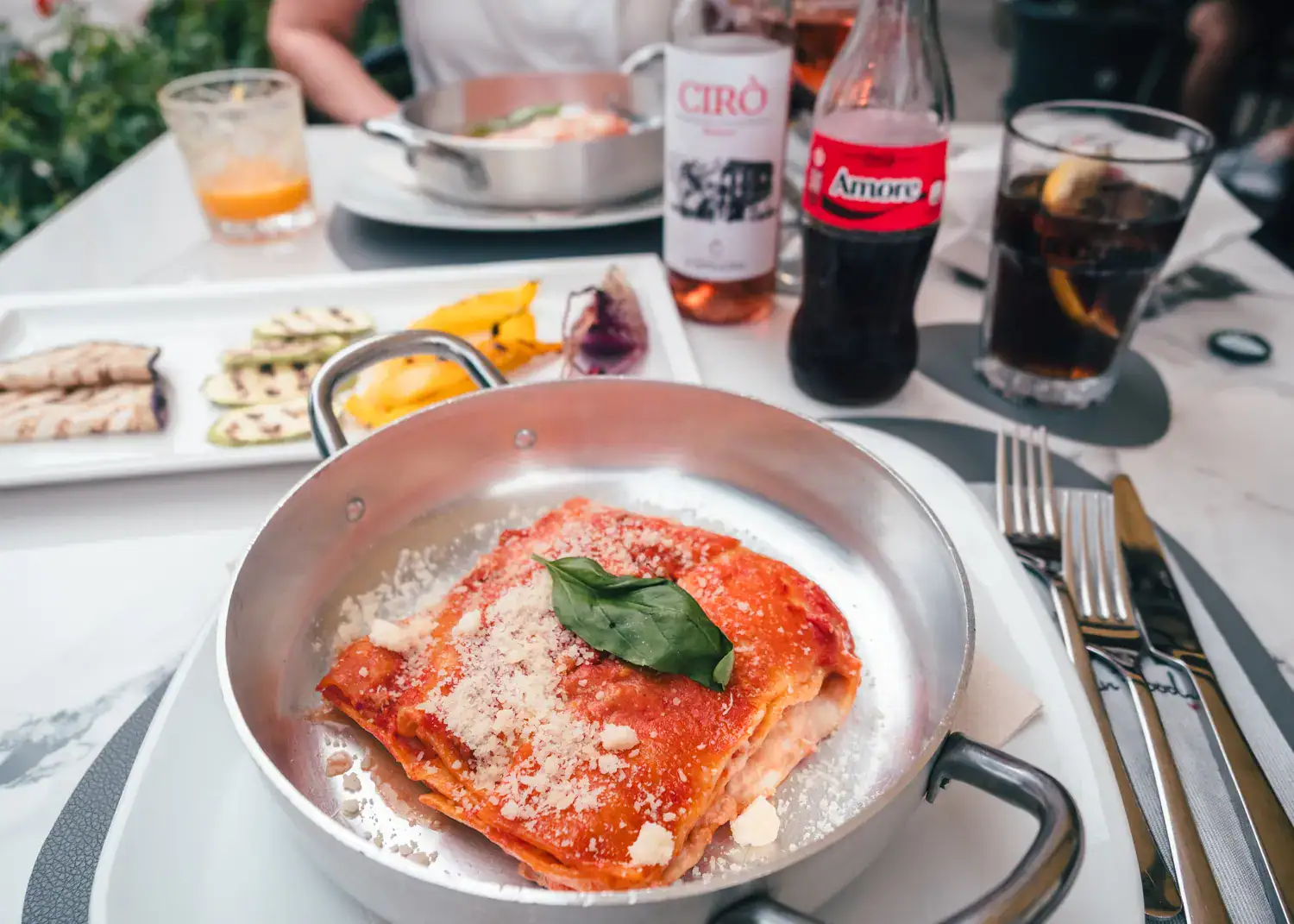 Silver dish with a piece of lasagne, white plate in the background with grilled vegetables, glass bottle of coke and a bottle of ros&eacute; on the table at Ristorante Tomate Calabrian Food in Tropea.
