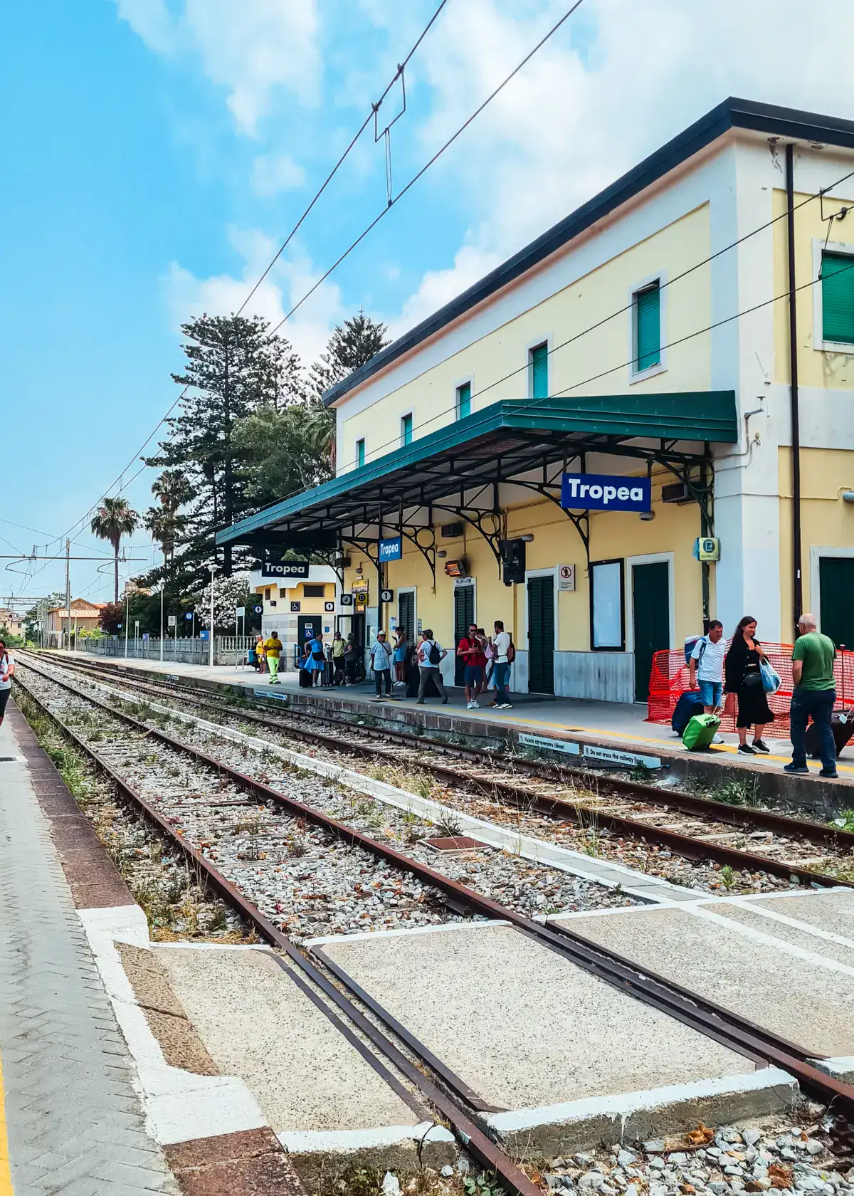 Tropea train stations in a yellow building with a green awning, people walking on the platform, transportation from Tropea to Lamezia Airport.