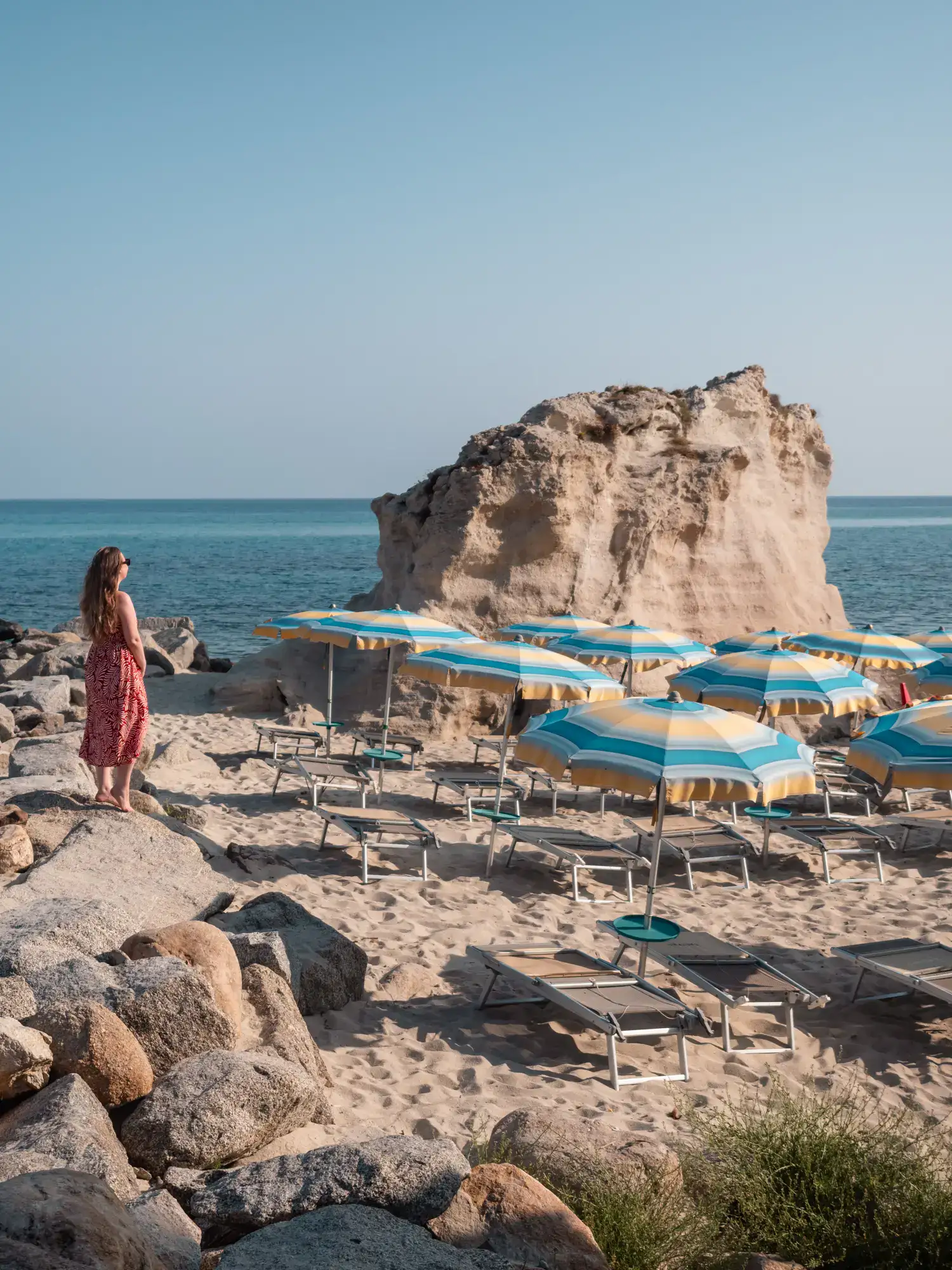 Woman wearing a red dress standing on rocks on the beach in Tropea, looking out over yellow and turquoise parasols, with a large sandstone rock in the background.