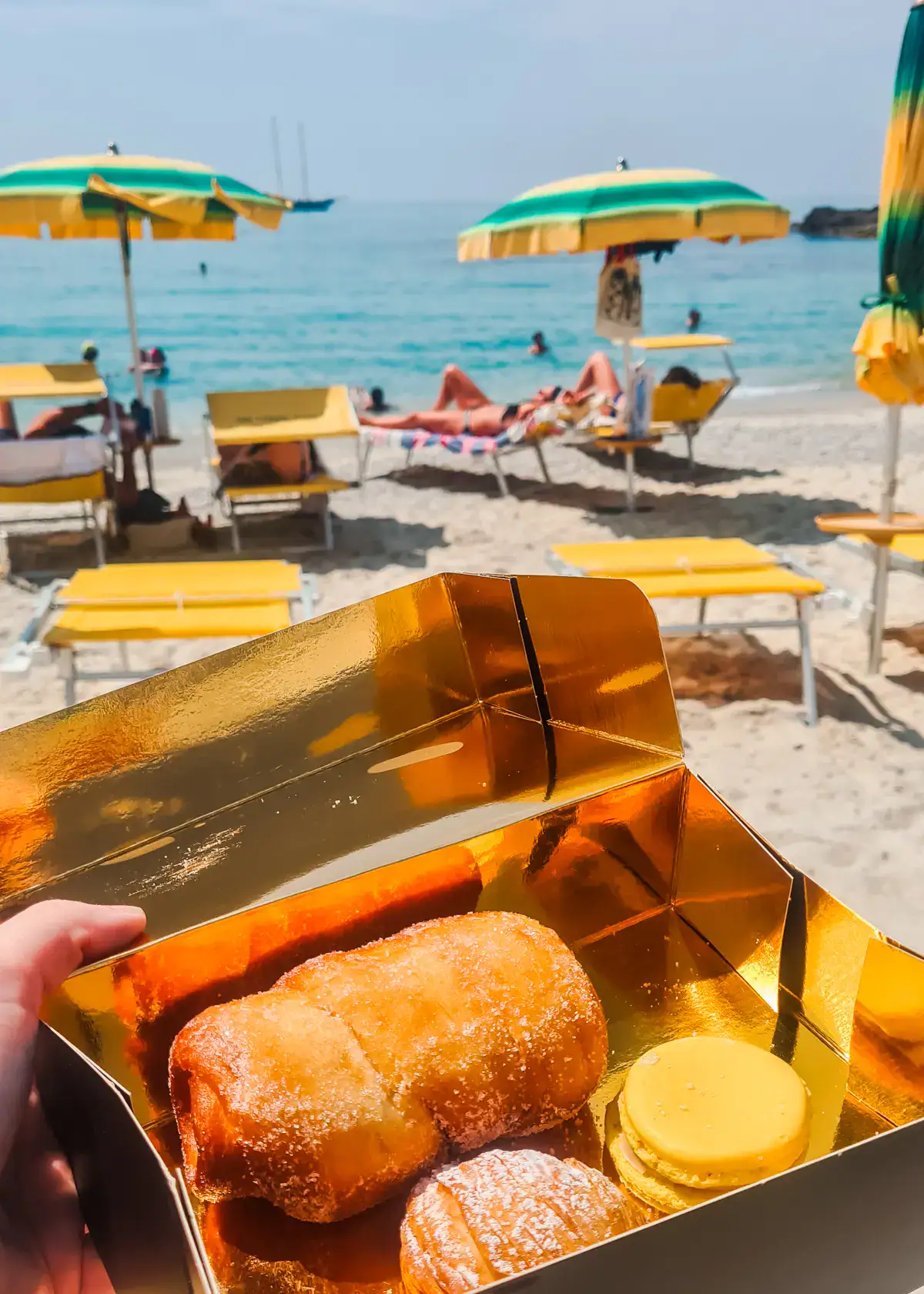 Gold box with three pastries from Premiata Pasticceria Tropea at the beach with yellow and green parasols and the blue ocean in the background in Tropea.