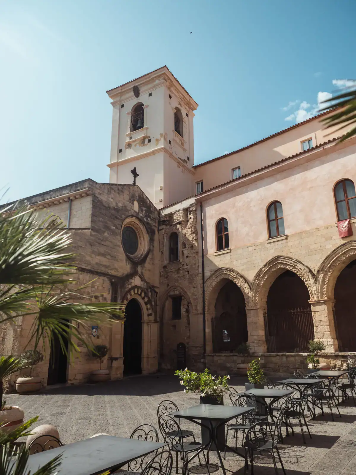 Sandstone colored Church of Saint Francis of Assisi with a tower and arches in a square with tables and chairs, one of the top things to see in Tropea.
