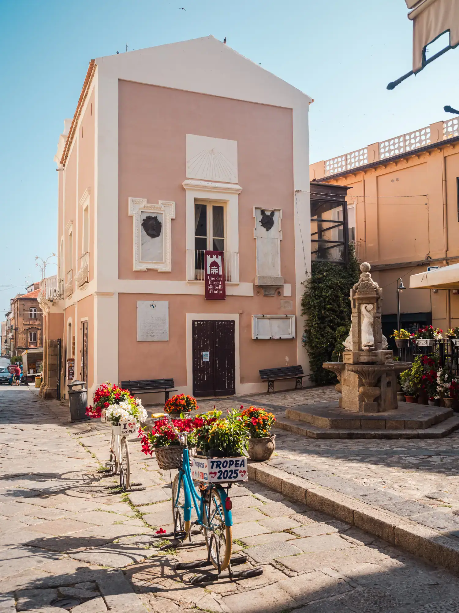 Light peach colored building with white details, stone fountain in a small square and two bikes with flowers in Tropea Old Town.