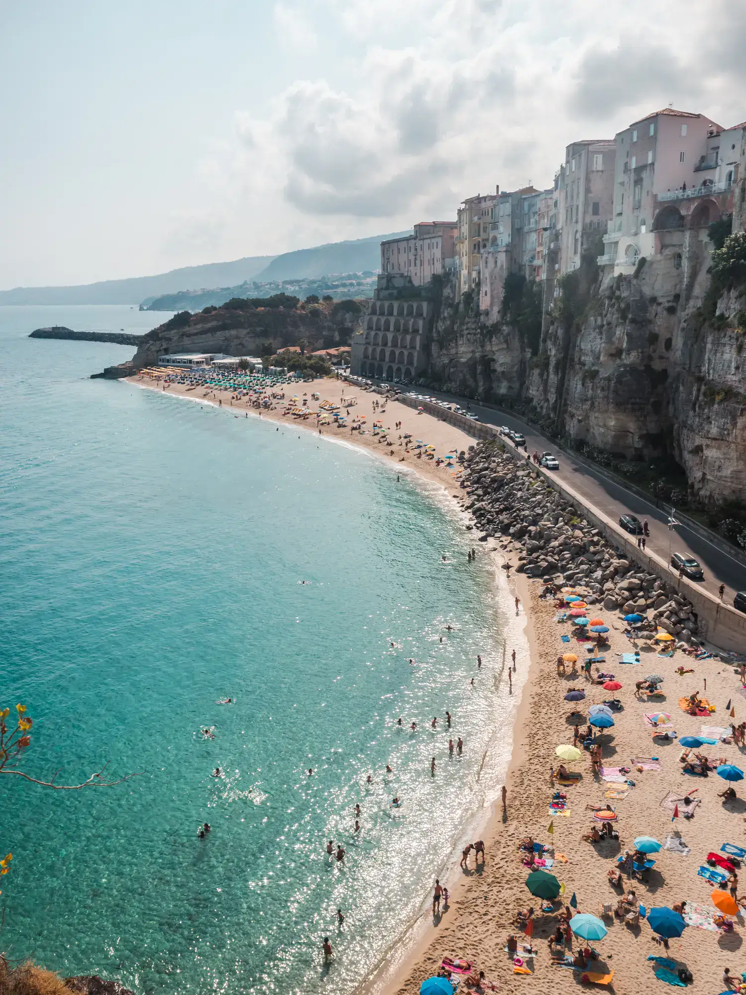 Beach with colorful parasols and people swimming in the turquoise ocean with Tropea Old Town built on a cliff on the right, Tropea Italy.