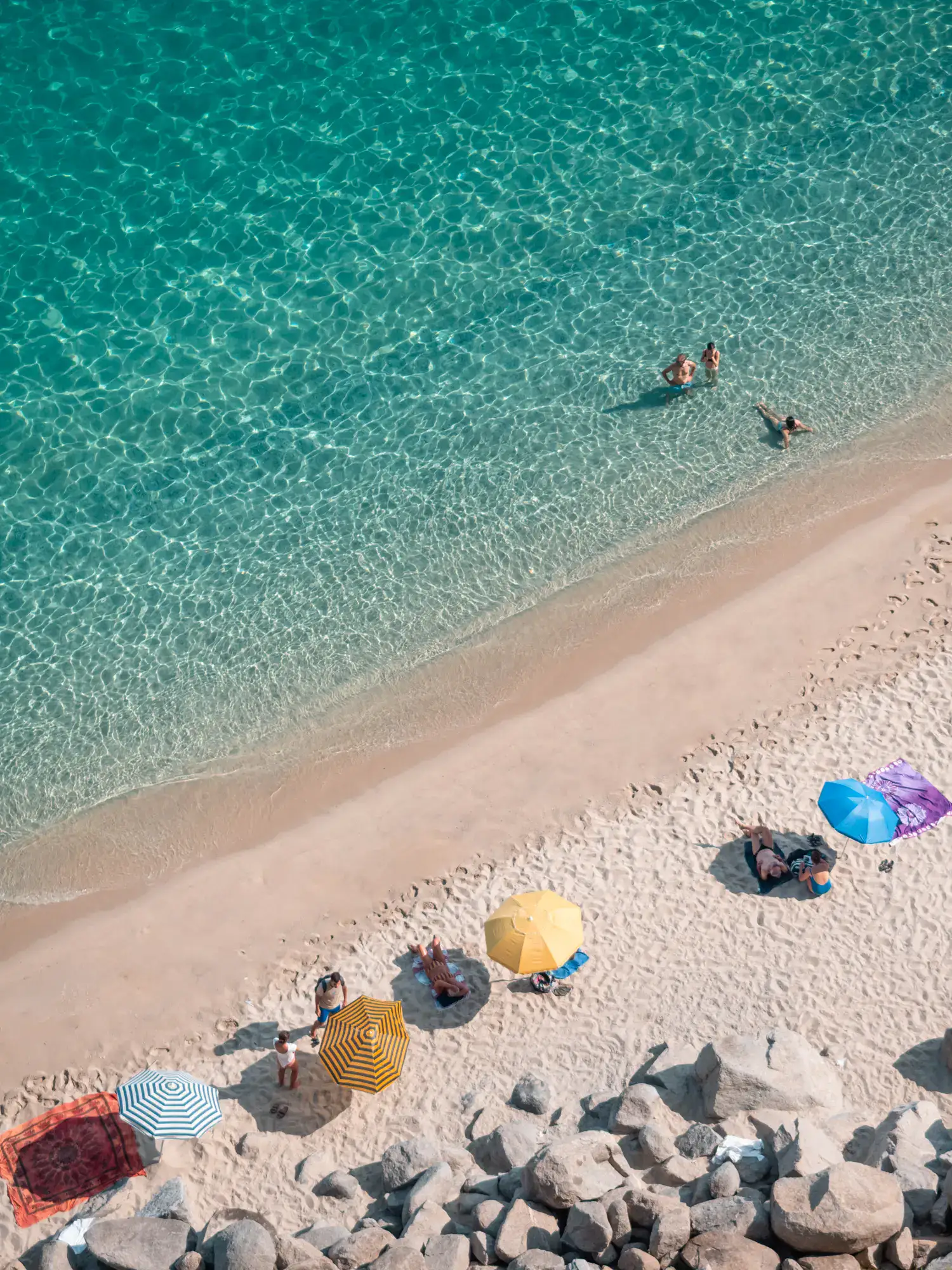 View from above of turquoise ocean, light beige sand, three people in the water, six colorful umbrellas on Beach in Tropea.