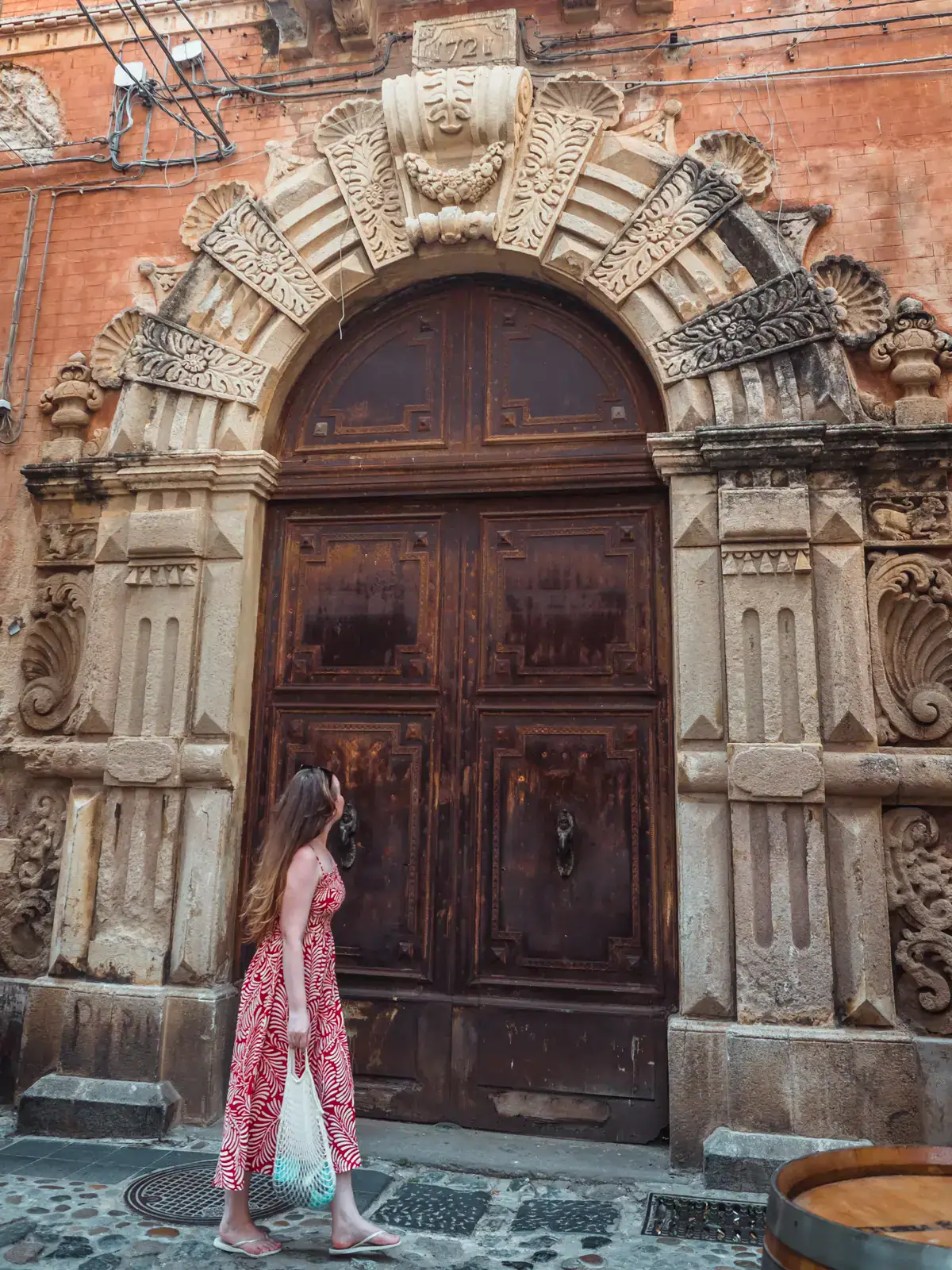 Woman wearing a red dress, holding a white bag, walking in front of a large brown arched door, with an intricate, old door frame in Tropea Old Town.