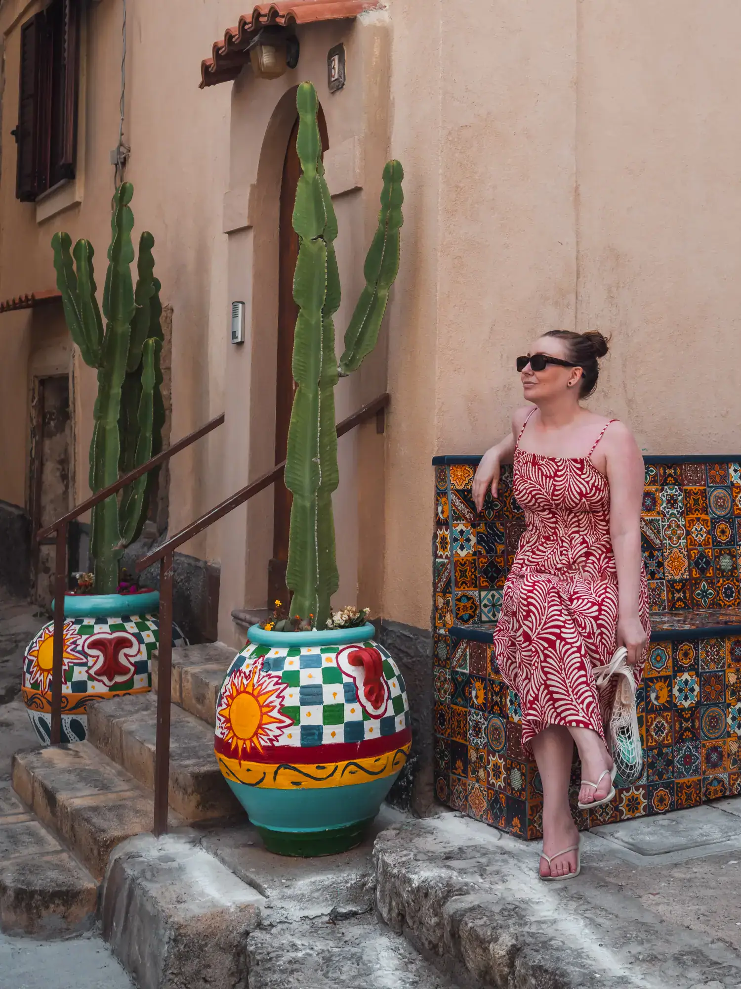 Woman wearing a red scrappy dress, hair in a bun and sunglasses, sitting on a tiles bench, with two large cacti in colorful pots, at Frida Mexican Restaurant in Tropea, Italy.