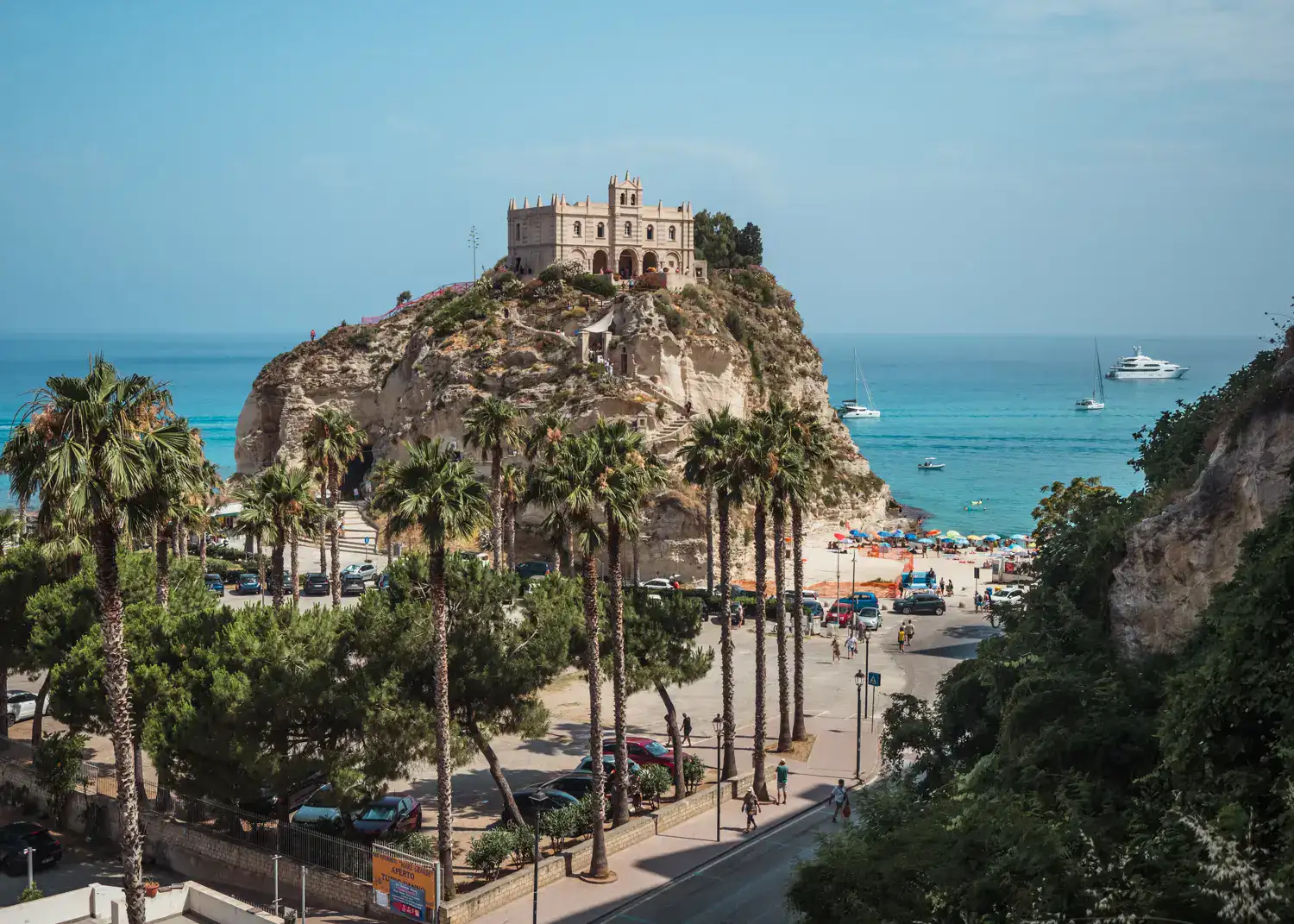 Santuario Santa Maria dell'Isola on top of a cliff by the ocean, skinny palm trees in the foreground, seen from one of the best viewpoints in Tropea Old Town. One of the top summer destinations in Europe.