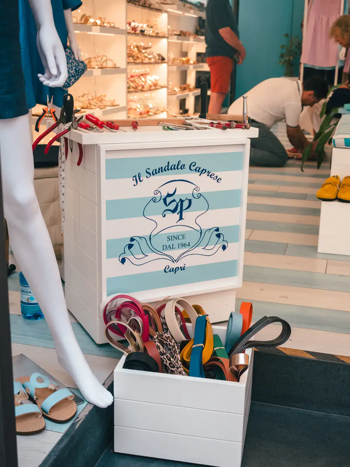 White box of leather sandals in front of a white and light blue striped counter and a white mannequin leg at a custom sandal/shoe shop in Tropea.