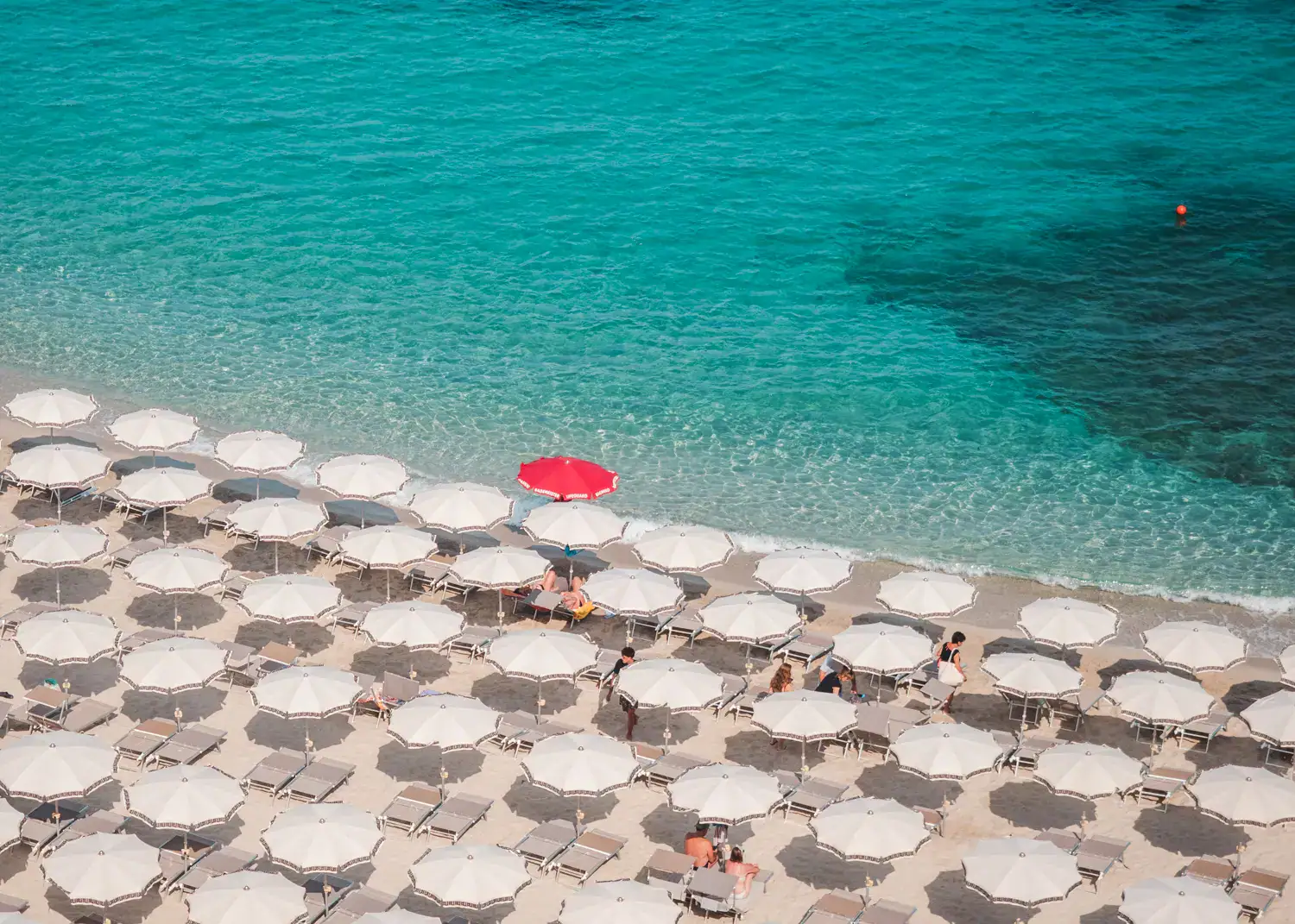 Rows of white parasols, with one red parasol in the middle by the turquoise ocean at Spiaggia a Linguata seen from above in Tropea.