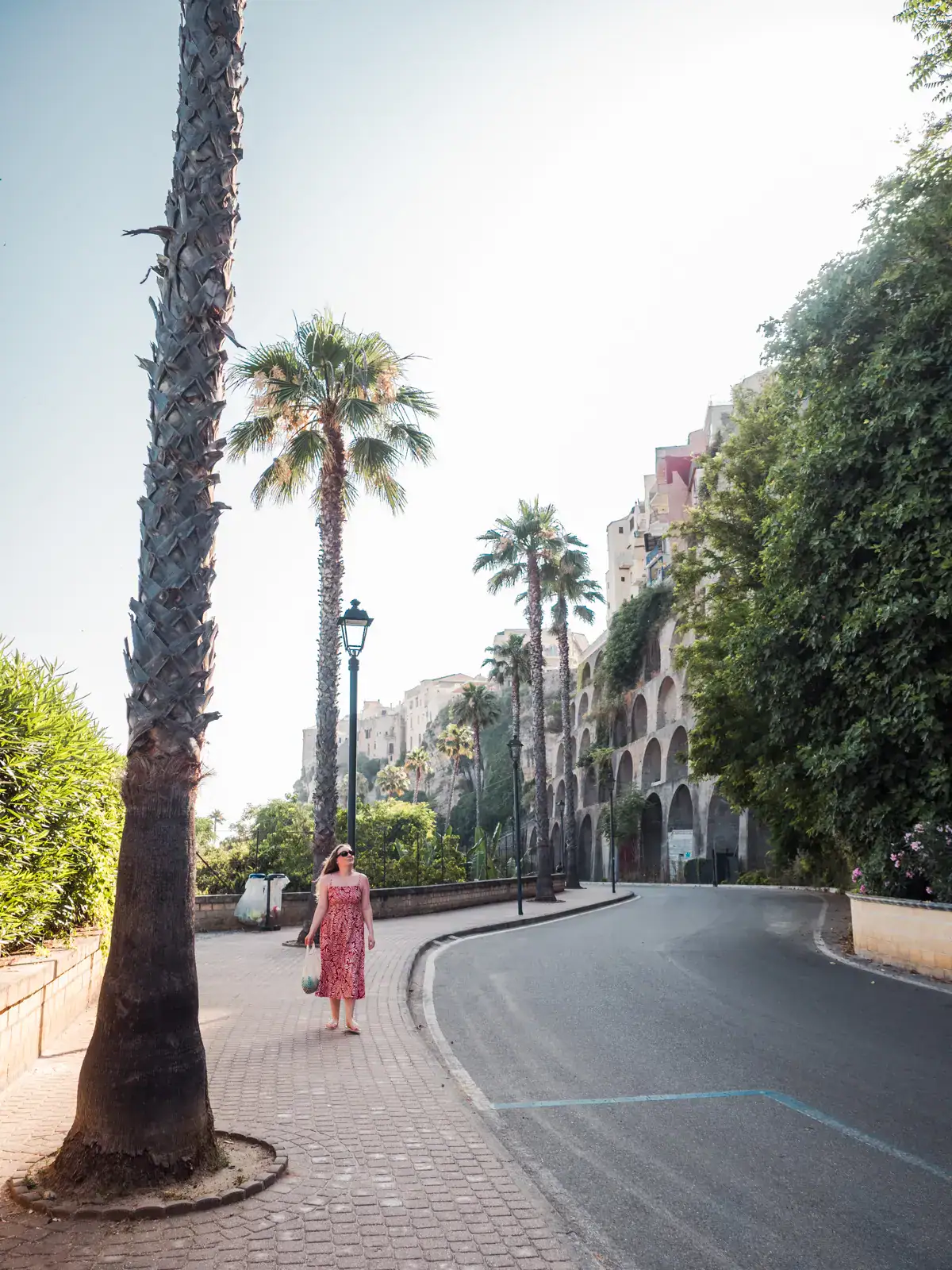 Woman wearing a red dress walking along a street lined with palm trees, next to a cliff in Tropea, Italy.