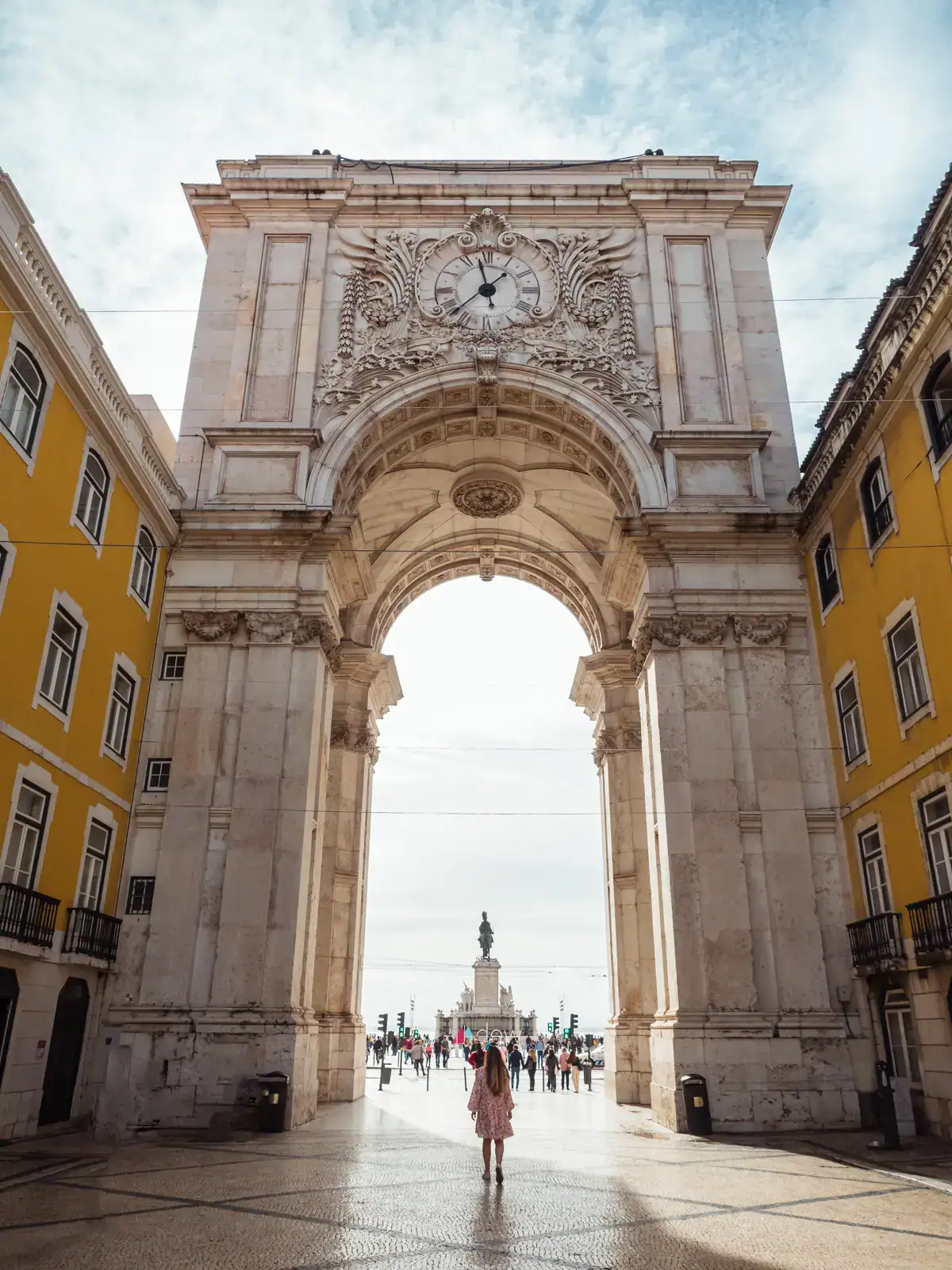 Woman walking under a tall, ornate arch with yellow buildings on both sides, sun shining into the arch in Lisbon, one of the best cities to visit in October.