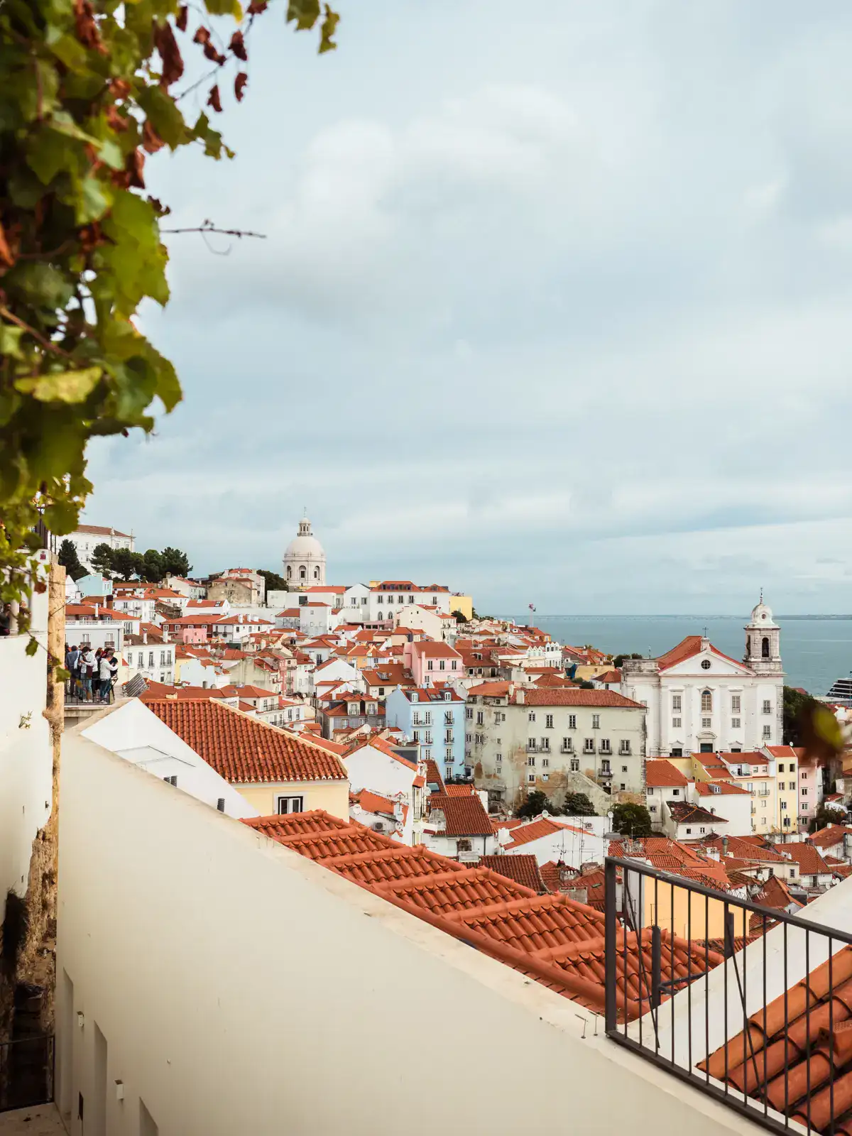 View across orange colored rooftops, pastel colored buildings and churches by the ocean from Miradouro de Santa Luzia in Lisbon, one of the best European cities to visit in October.