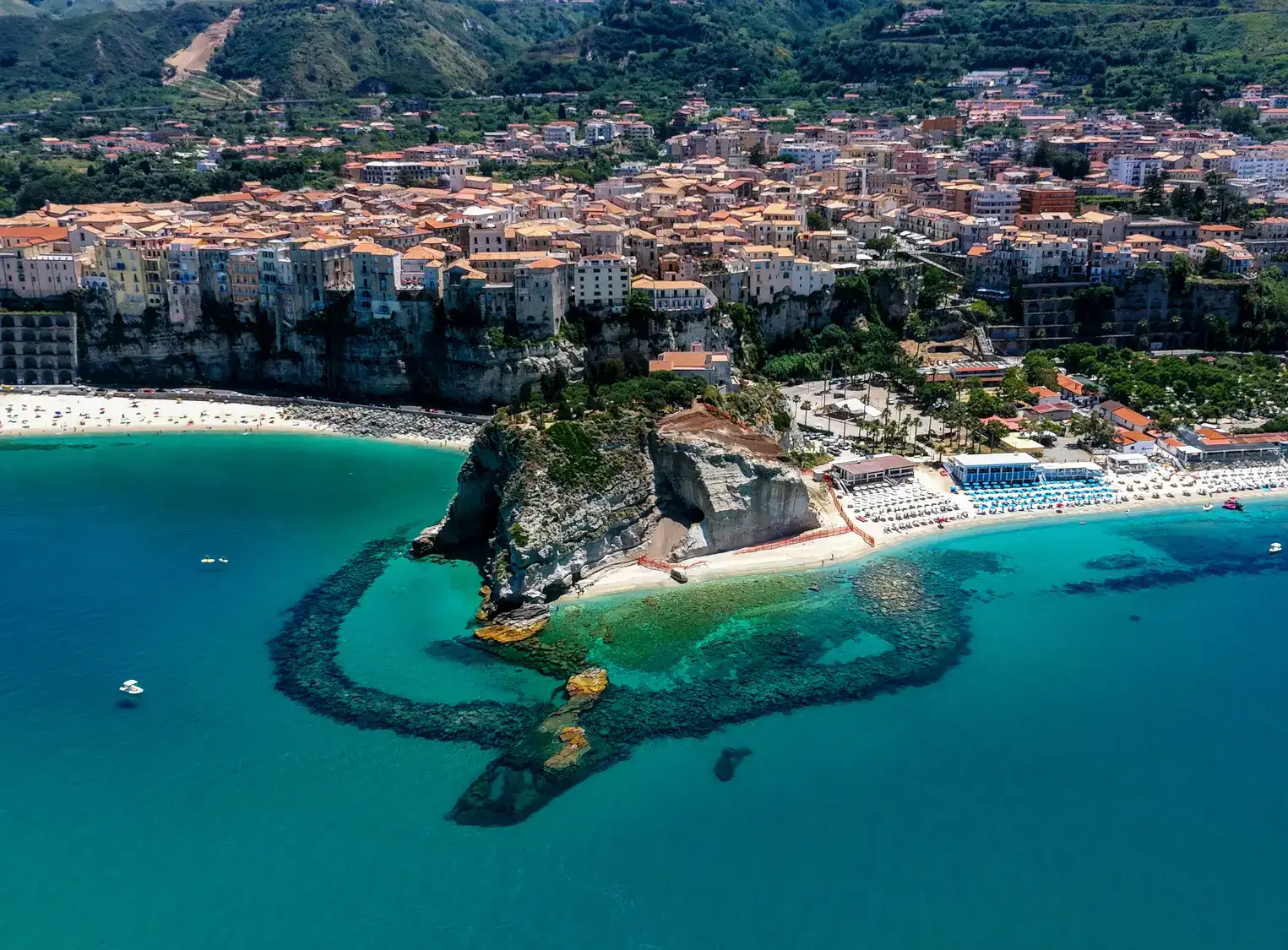 Aerial view of Tropea's famous rock, with beaches on both sides, including Spiaggia di Tropea, turquoise ocean and the old town on a cliff in the background.