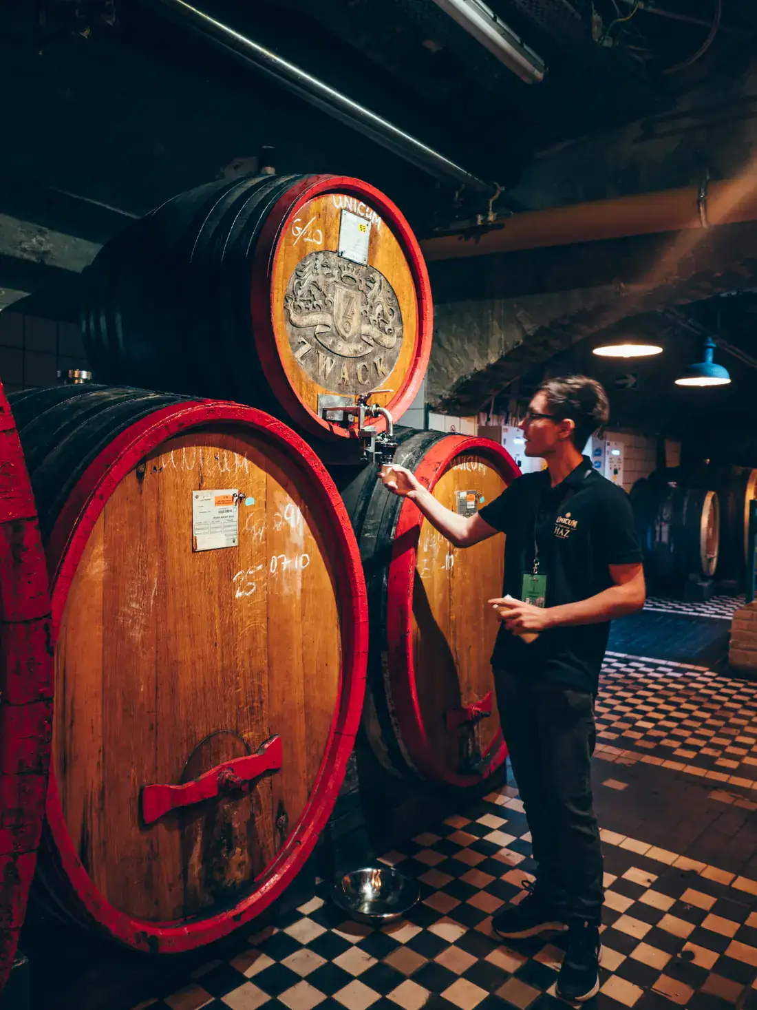 Our guide, tapping Unicum from a wooden barrel with the Unicum logo on top of two larger barrels, during a premium tour of the Factory and Museum in Budapest.
