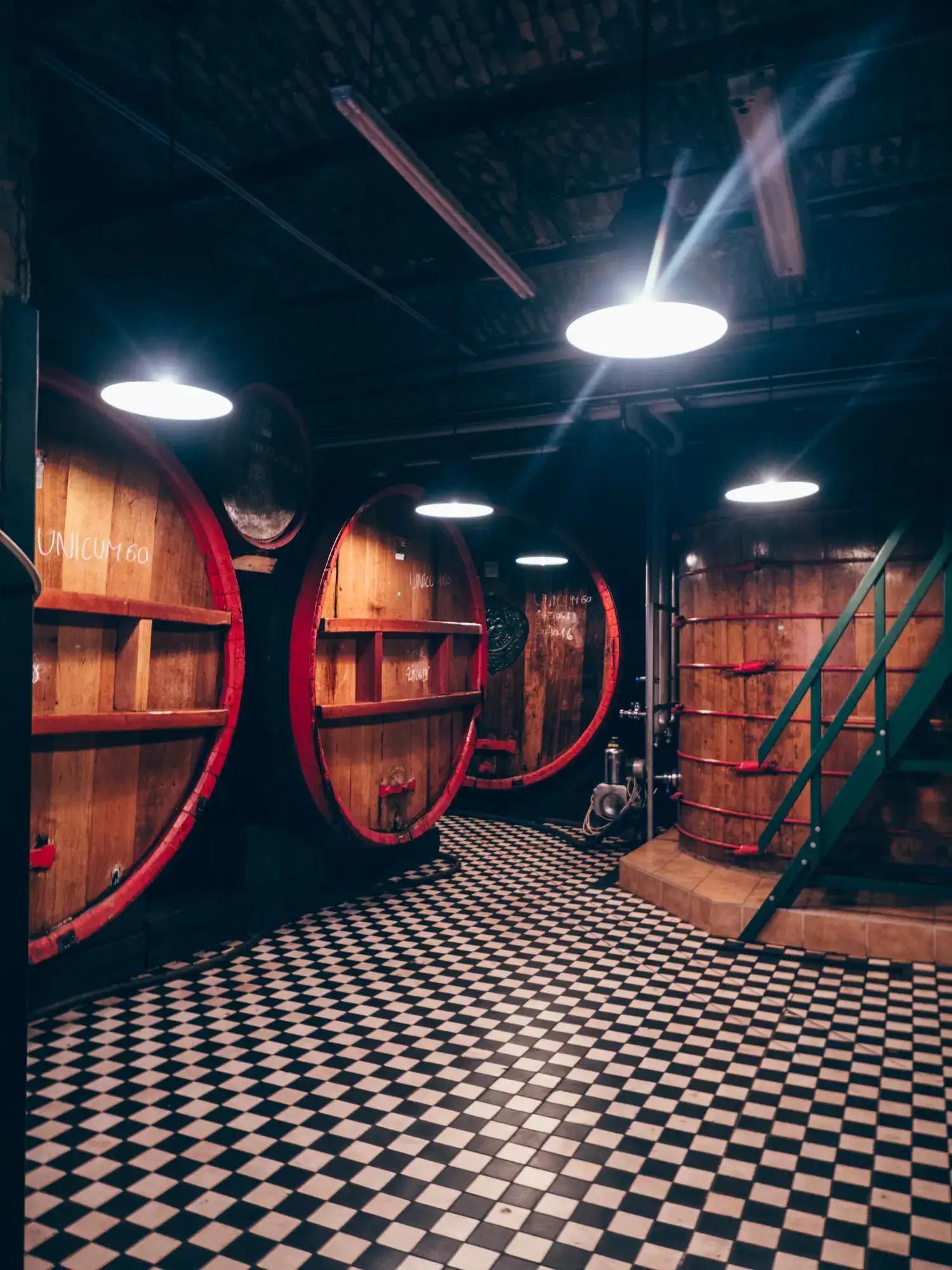 One of the rooms in the Zwack Unicum Factory cellar, with small, white and black checkered floor tiles, large wooden barrels with red trim, green metal staircase and round lights in the ceiling.
