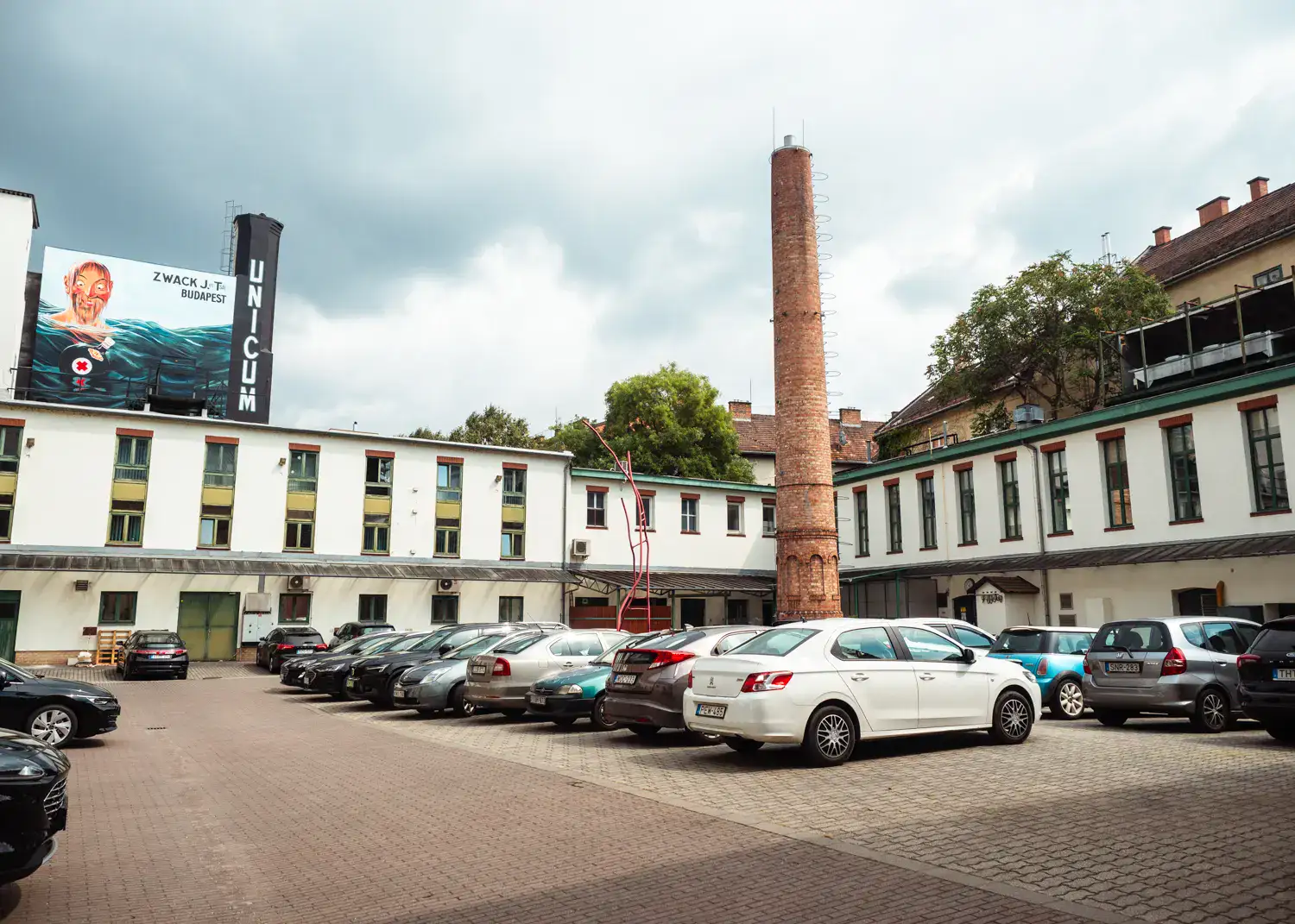 View inside courtyard of the Zwack Unicum Factory, with cars parked in the middle, white factory building surround it, brown brick chimney and a large Zwack Unicum poster in Budapets.